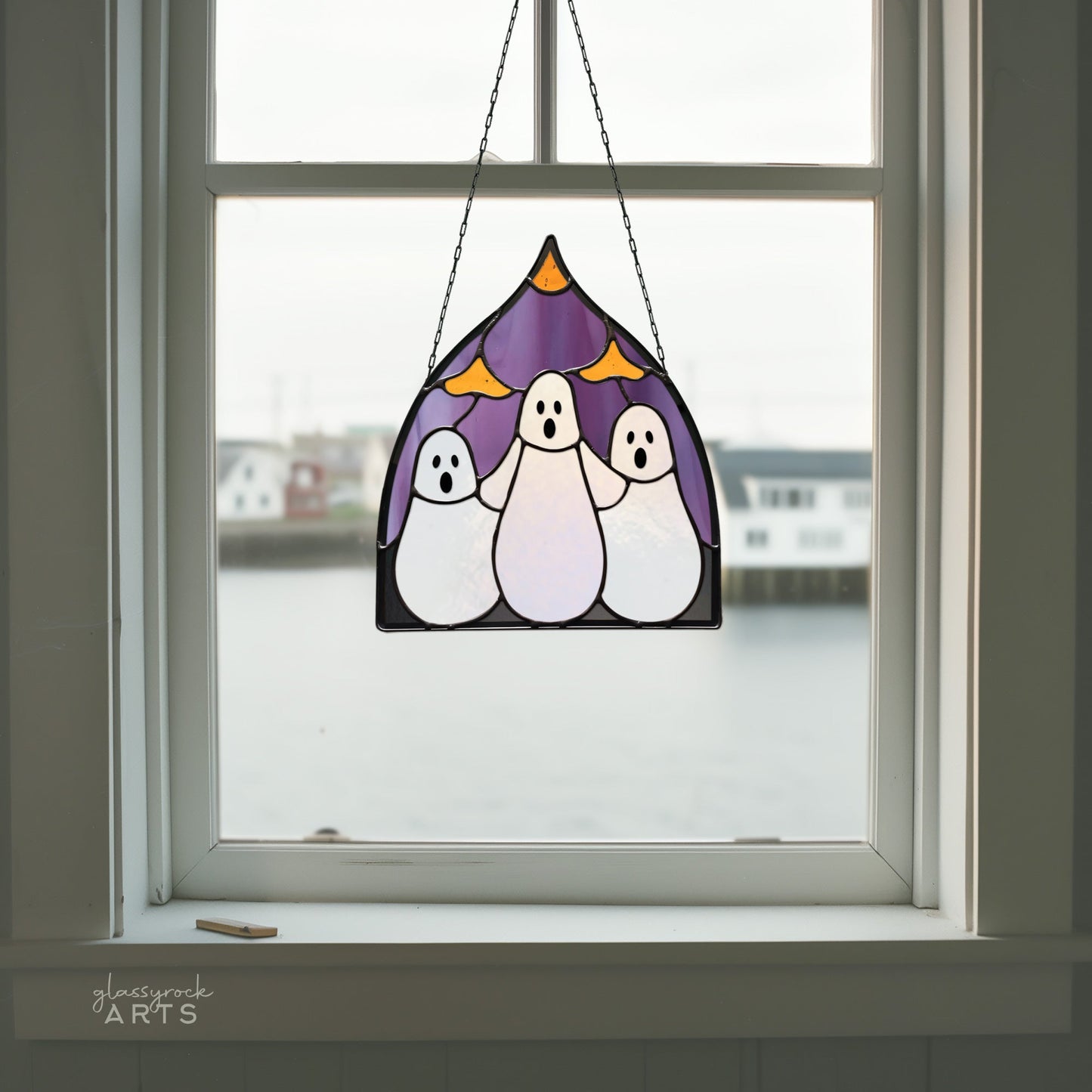 A Three Ghosts Halloween Stained Glass Pattern features stained glass suncatcher ghost figures with purple and yellow accents. The window scene outside is blurred, showing water and a building. Download the digital pattern to craft your own eerie masterpiece.