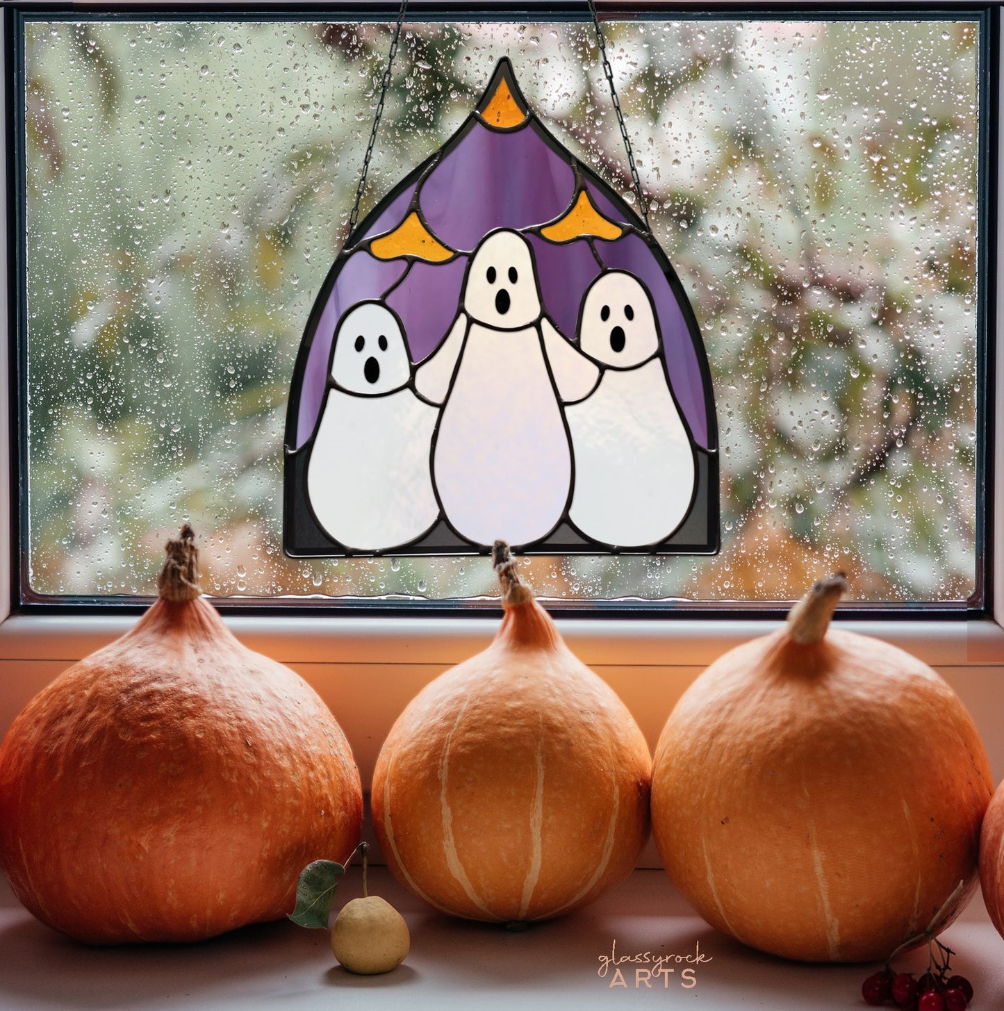 The Three Ghosts Halloween Stained Glass Pattern hangs in a rain-specked window with three pumpkins, a green apple, and red berries on the sill, forming an enchanting autumnal scene.
