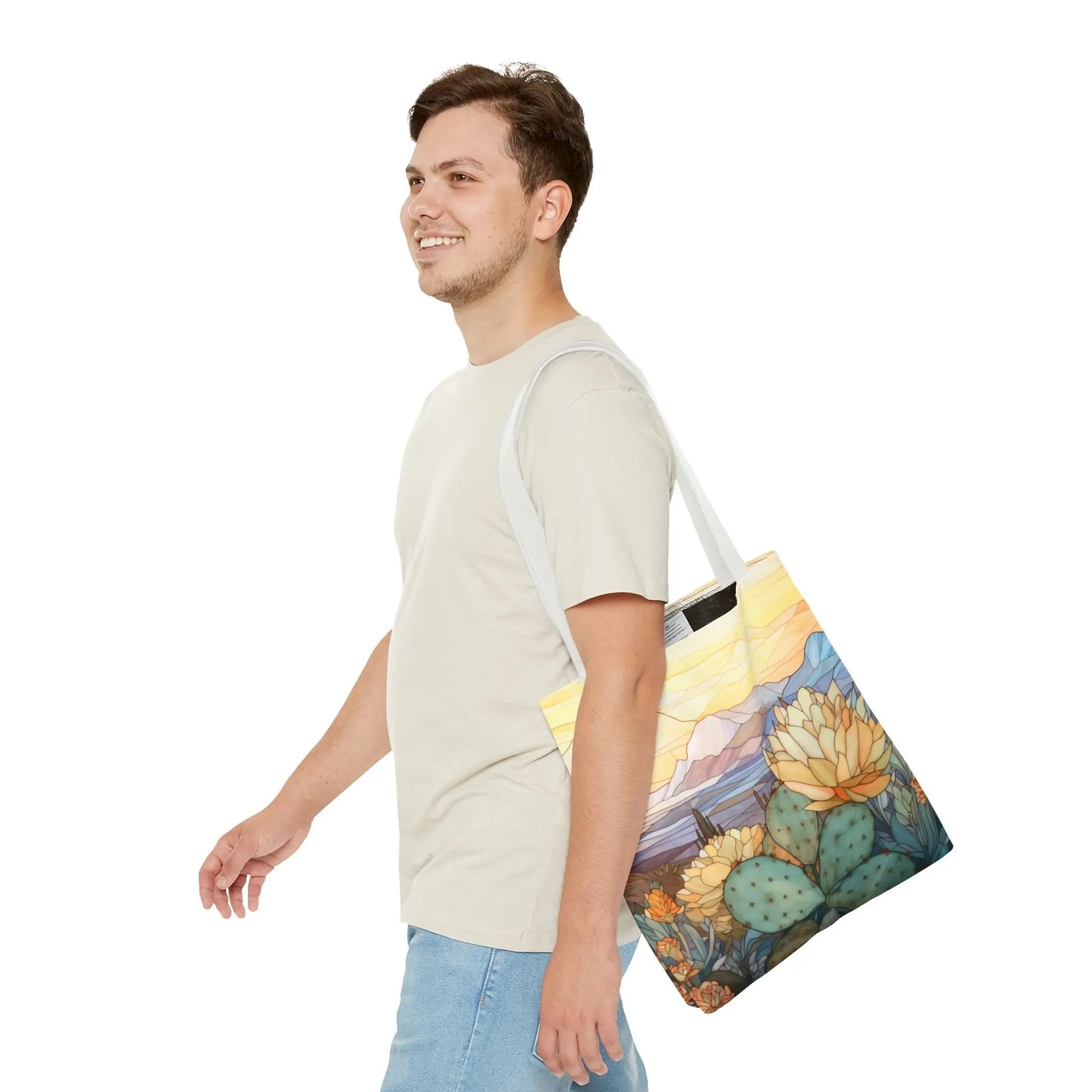A person with short dark hair in a beige t-shirt and blue jeans smiles as they walk, carrying the Stained Glass Desert Cactus Tote Bag made from durable polyester, featuring an original design of cacti and mountains.