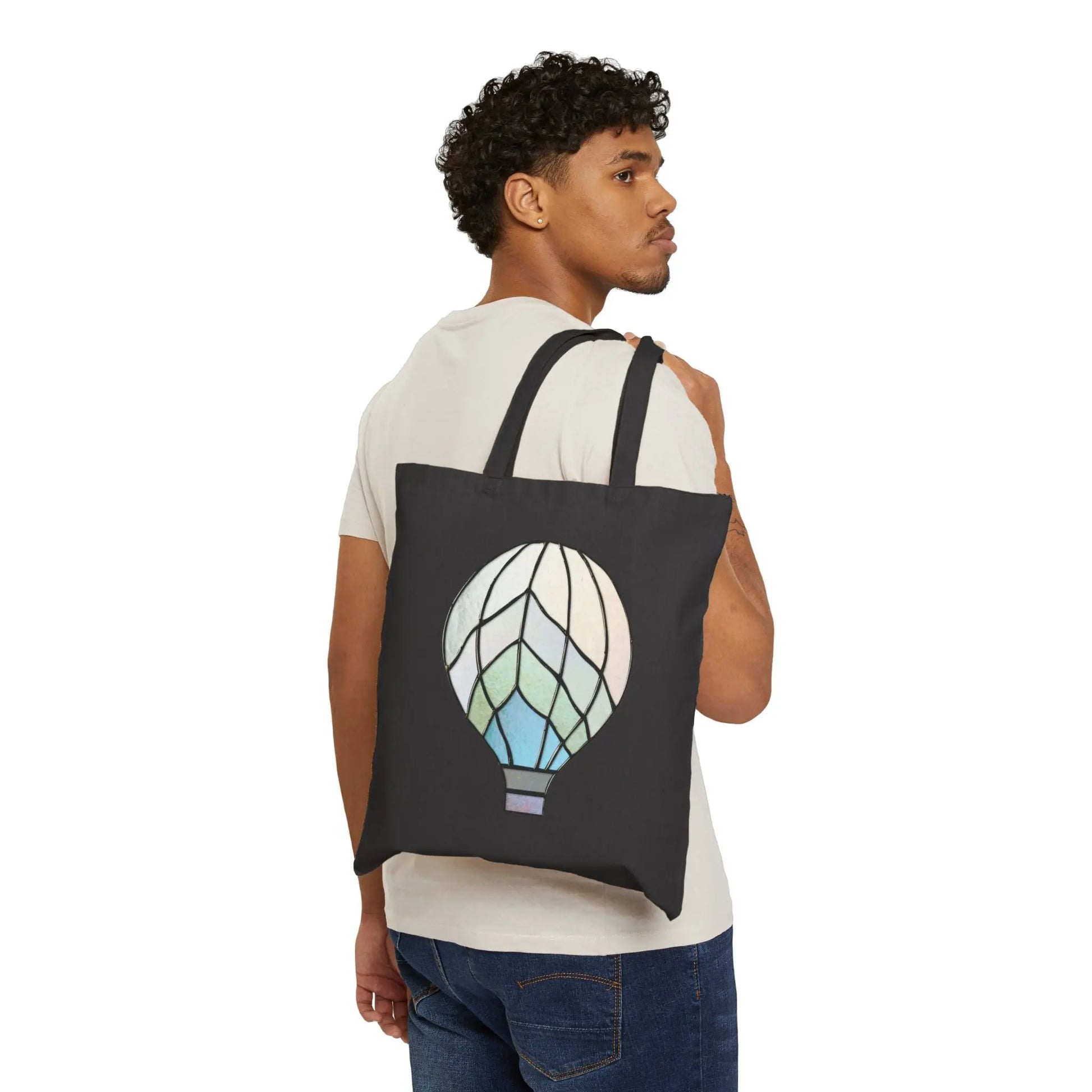 A curly-haired person wears a light t-shirt and blue jeans, holding the Stained Glass Hot Air Balloon Cotton Canvas Tote Bag on their shoulder. The black tote is 100% cotton with an abstract hot air balloon design in a pastel stained glass pattern.
