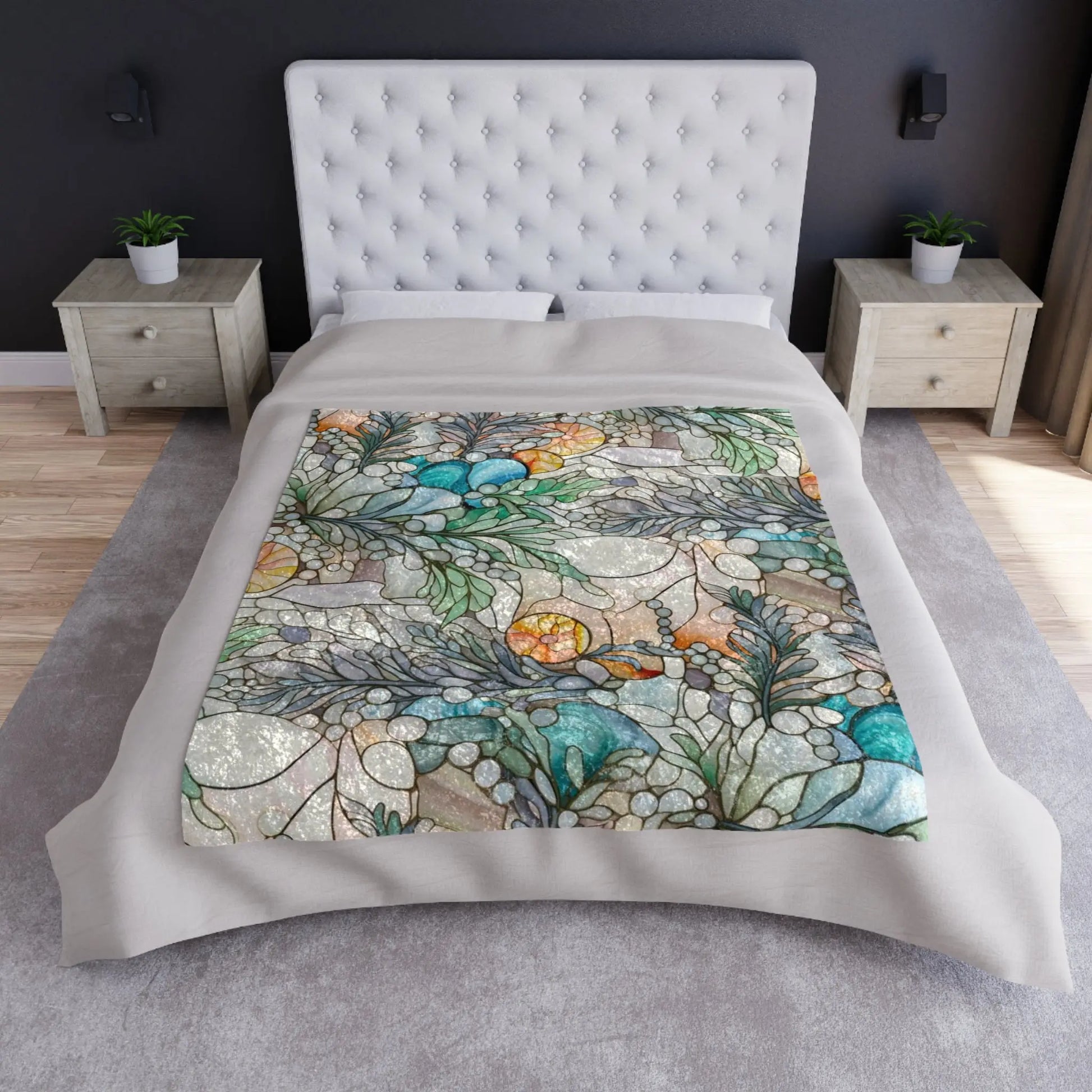 A bed with a tufted white headboard features the Stained Glass Rainbow Ferns Forest Crushed Velvet Blanket, showcasing swirling blues, greens, and oranges. It stands in a room with dark walls, light wooden side tables, and small plants on each table.