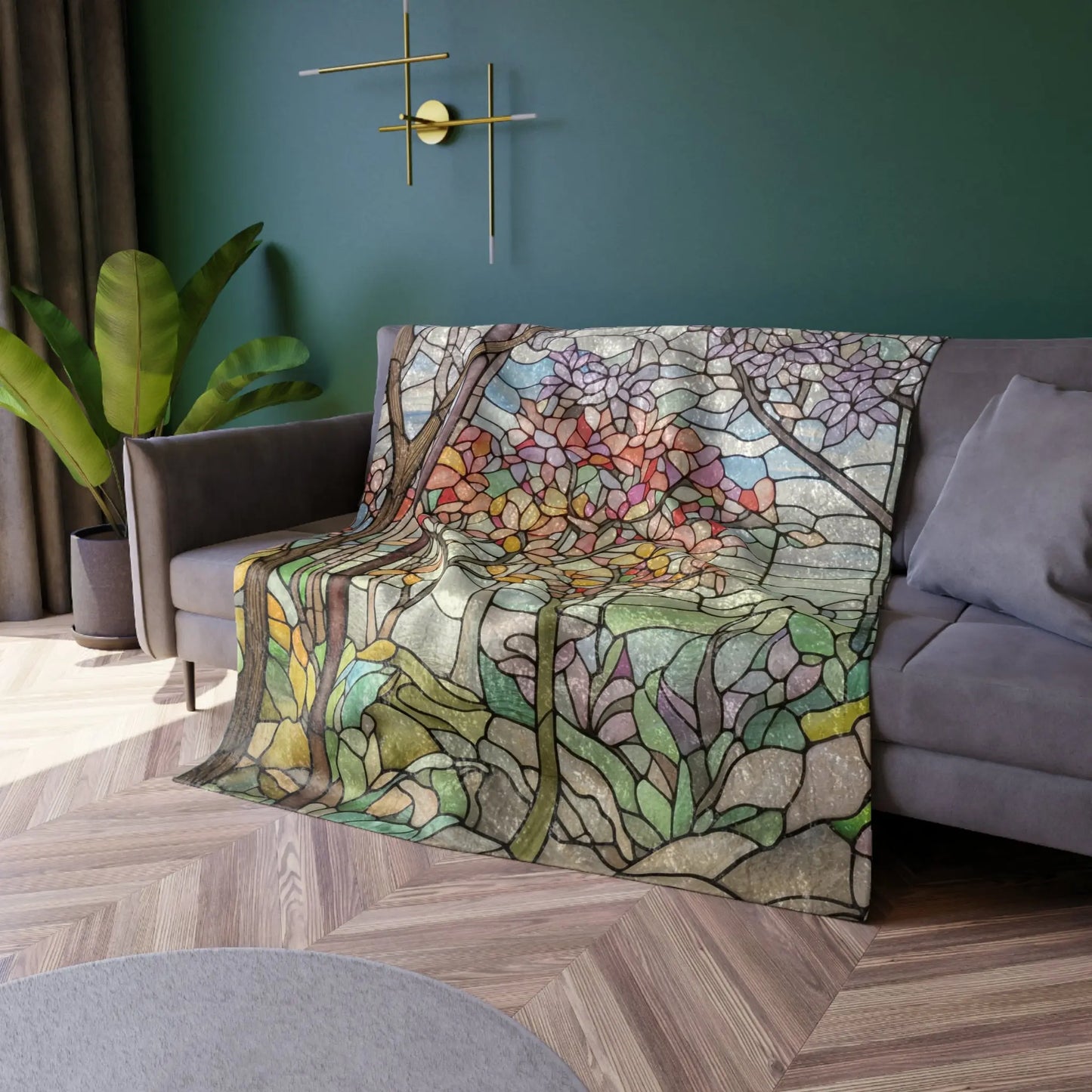 A Stained Glass Springtime Forest Crushed Velvet Blanket in a 50x60 size, featuring colorful floral patterns, is draped over a gray sofa. The room showcases a green wall, modern gold wall sconce, potted plant, and chevron wood flooring.