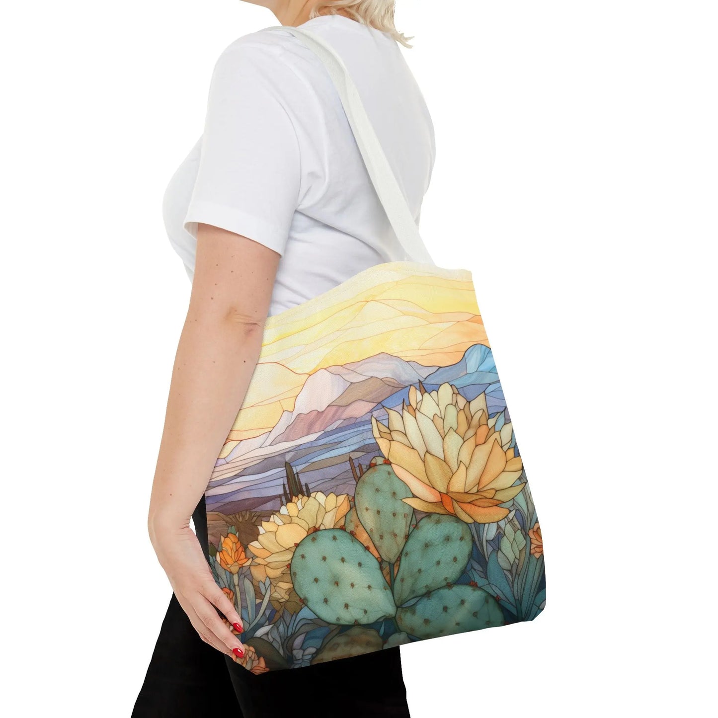 A person in a white T-shirt carries the Stained Glass Desert Cactus Tote Bag, featuring a colorful desert scene with cacti, blooming flowers, mountains, and a vibrant sky on durable polyester material.