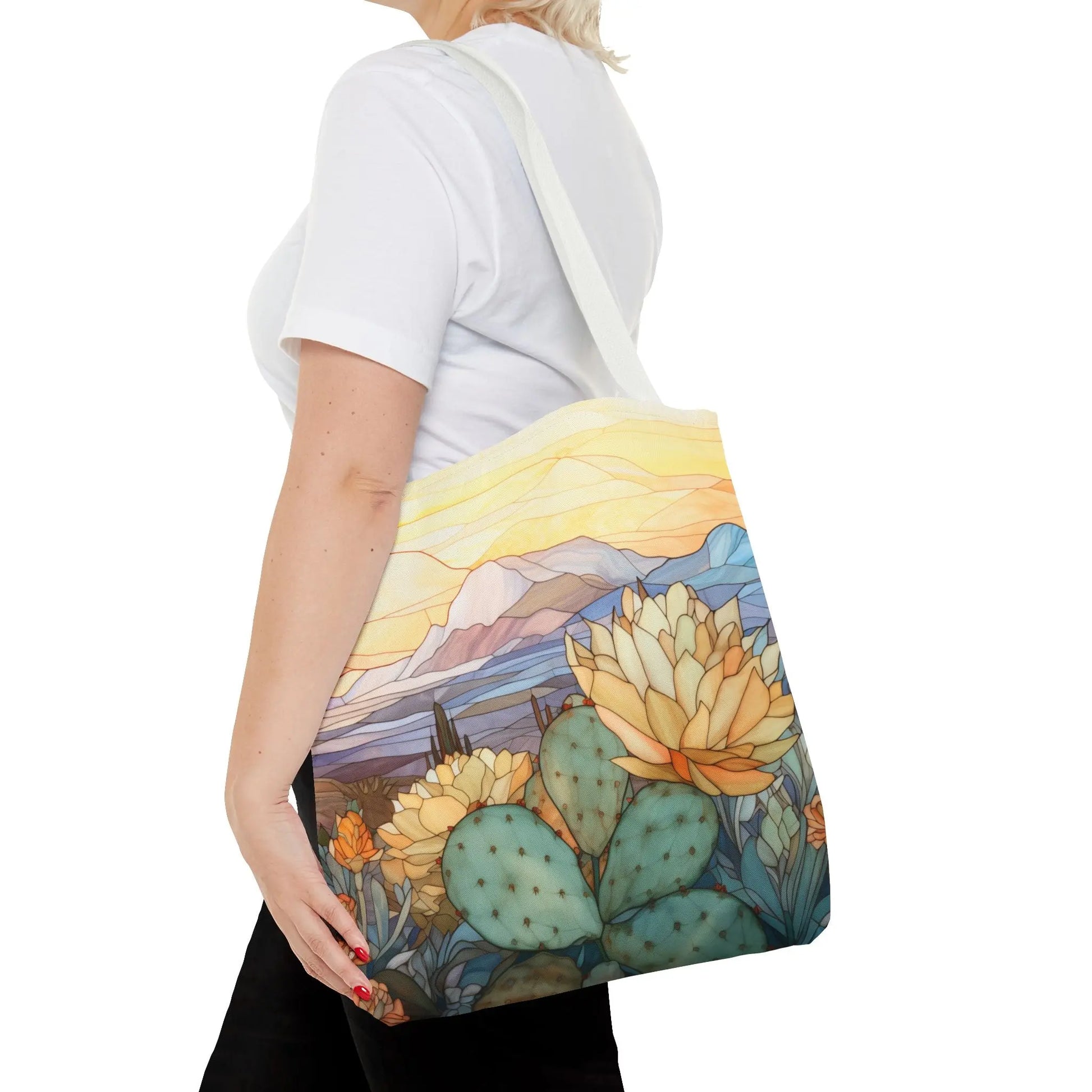 A person in a white T-shirt carries the Stained Glass Desert Cactus Tote Bag, featuring a colorful desert scene with cacti, blooming flowers, mountains, and a vibrant sky on durable polyester material.