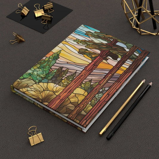 A Stained Glass Sequoia Forest Hardcover Journal with a sunset and trees design rests on a textured surface, surrounded by gold and black pencils, gold binder clips, and a geometric decor piece, making this custom creation both functional and artistically delightful.