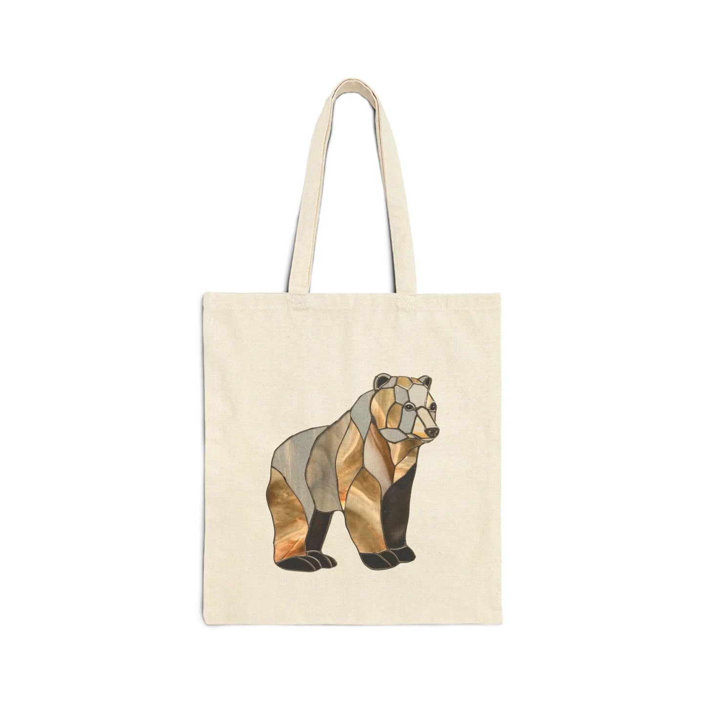 The Stained Glass Bear Cotton Canvas Tote Bag features a beige canvas with a stylized bear design in geometric brown, black, and gold shades inspired by Noelle Barnes stained glass art. It includes two handles and is displayed against a white background.