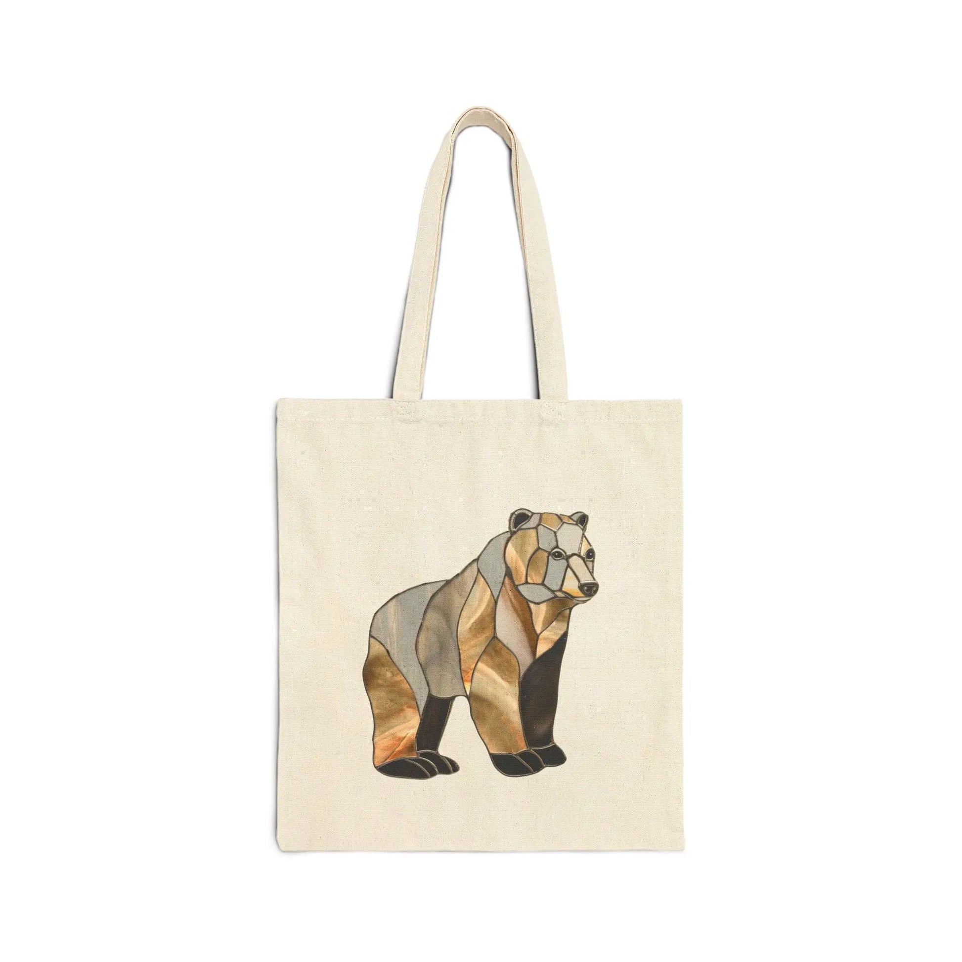 The Stained Glass Bear Cotton Canvas Tote Bag features a beige canvas with a stylized bear design in geometric brown, black, and gold shades inspired by Noelle Barnes stained glass art. It includes two handles and is displayed against a white background.