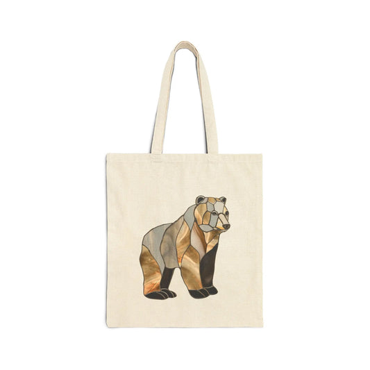 The Stained Glass Bear Cotton Canvas Tote Bag features a beige canvas with a stylized bear design in geometric brown, black, and gold shades inspired by Noelle Barnes stained glass art. It includes two handles and is displayed against a white background.