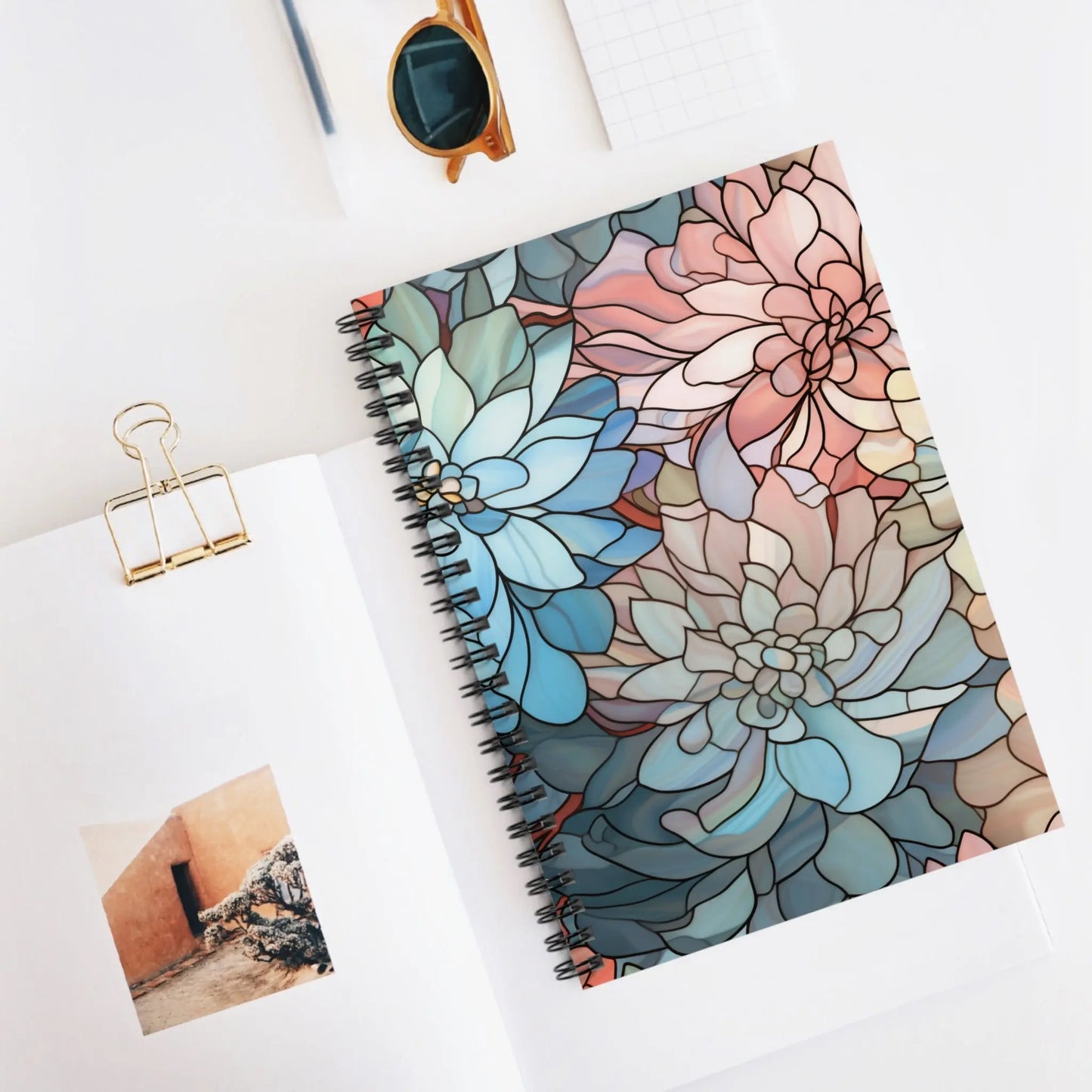 The Stained Glass Dark Flowers Spiral Notebook with a floral cover rests on an open magazine. Nearby are sunglasses, a blank notepad, and a paper clip holding a small photo, creating a bright and minimalistic setting—a perfect gift for stained glass lovers.