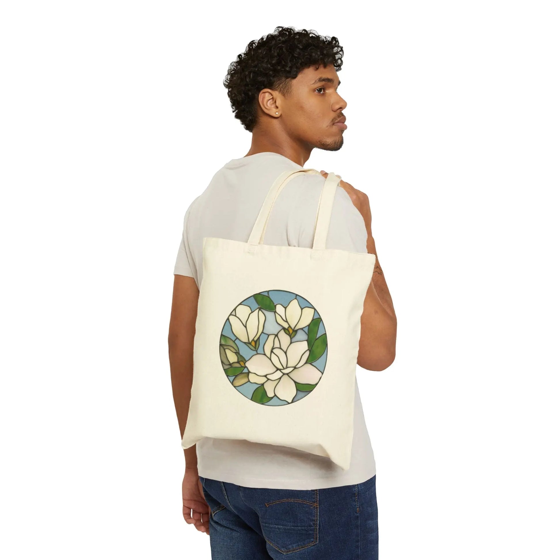A curly-haired person stands sideways wearing a light gray T-shirt and blue jeans, with the Stained Glass Magnolia Cotton Canvas Tote Bag over their shoulder—a cream-colored bag featuring a floral design with white flowers and green leaves. Perhaps its by Noelle Barnes.