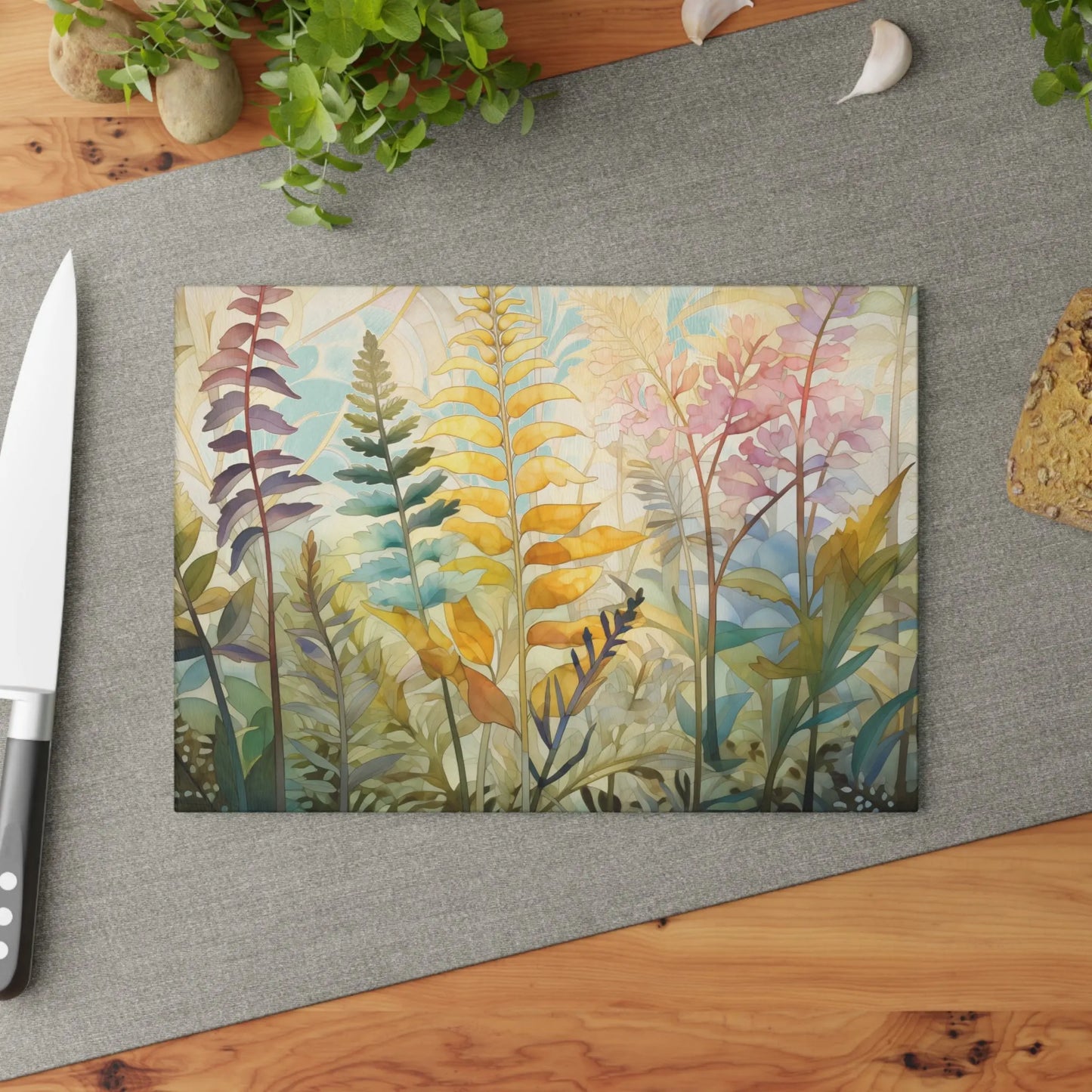 On a wooden table, the Stained Glass Pastel Ferns Glass Cutting Board displays colorful abstract leaves beside a knife. It sits on a gray fabric runner with green plant sprigs and garlic in the background.