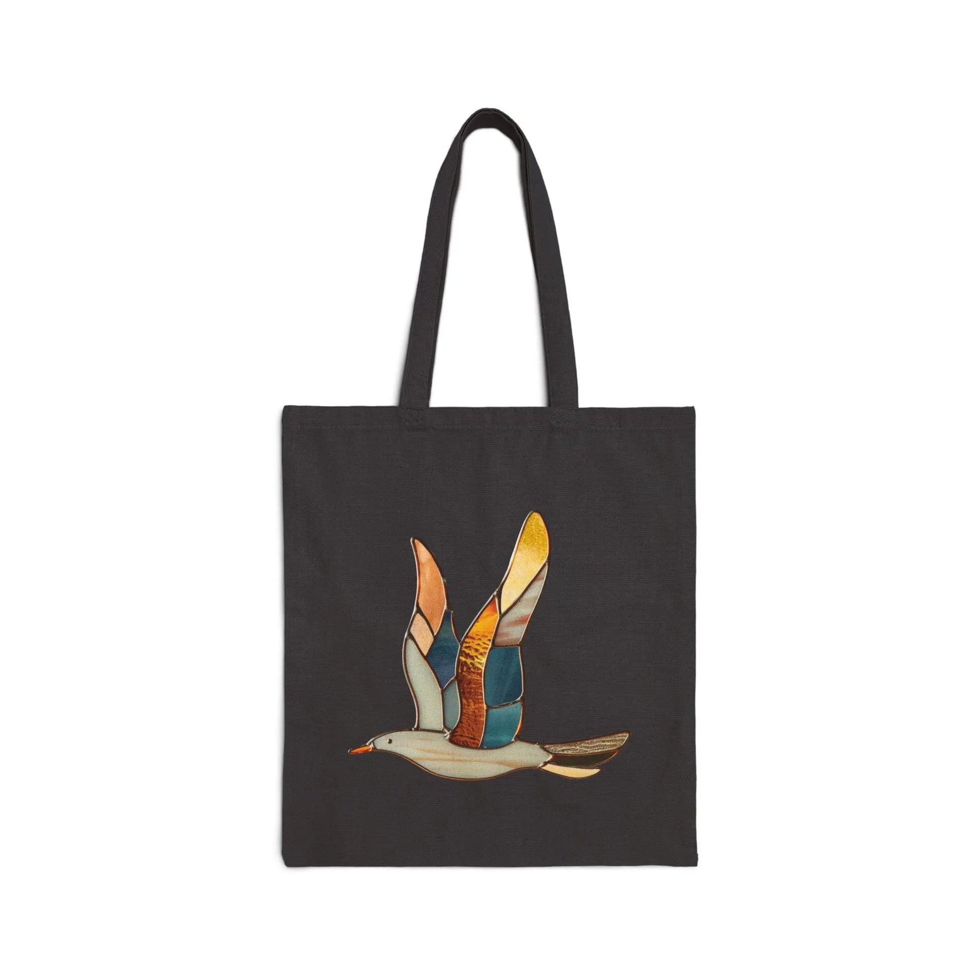 The Stained Glass Seagull Cotton Canvas Tote Bag is a black bag with a vibrant design showcasing a stylized seagull in flight, featuring wings with colorful patches of orange, yellow, and blue.