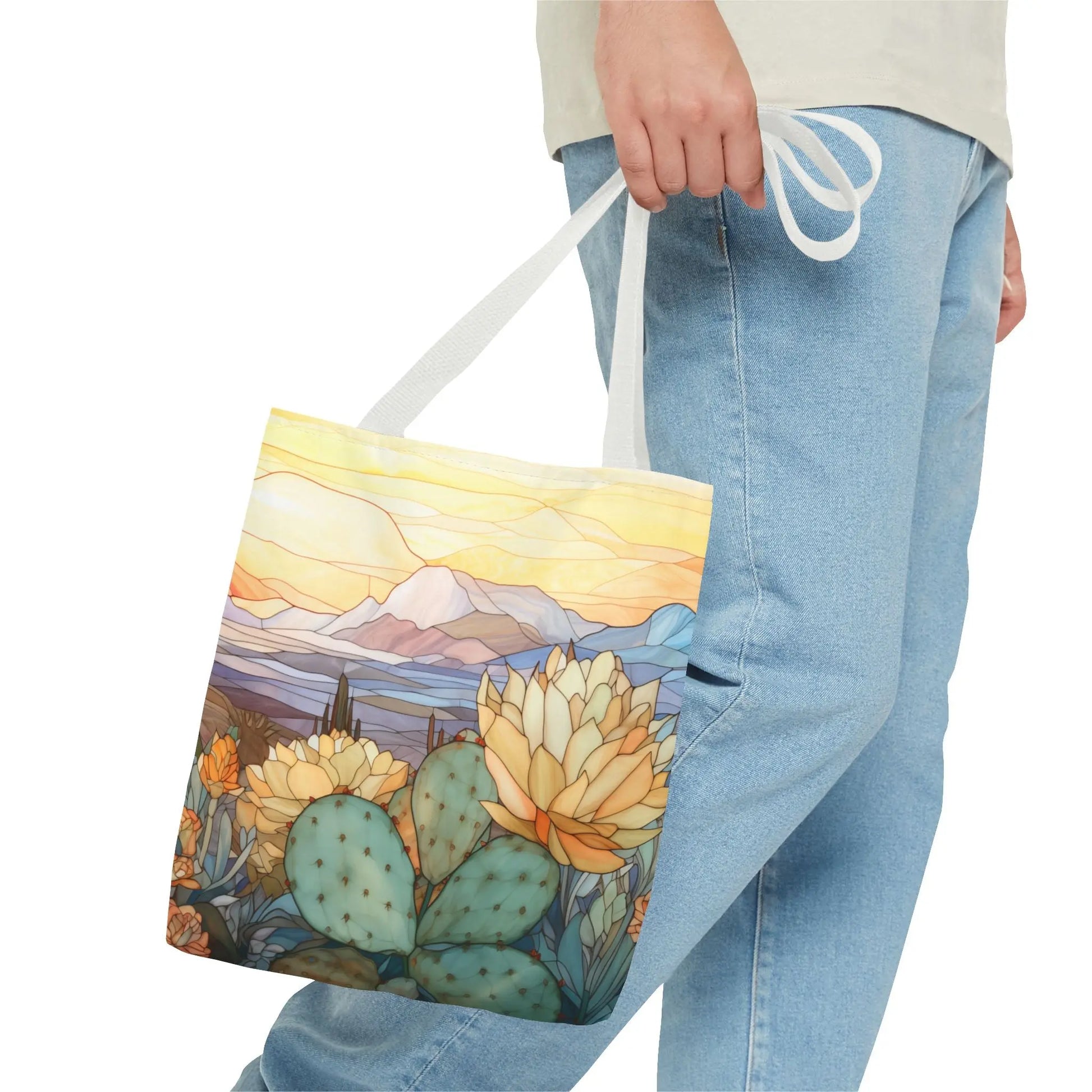 A person in light blue jeans holds a Stained Glass Desert Cactus Tote Bag, featuring an original artist design with cacti, flowers, mountains, and a sunrise or sunset. The colorful bag is crafted from durable polyester and available in 3 sizes.