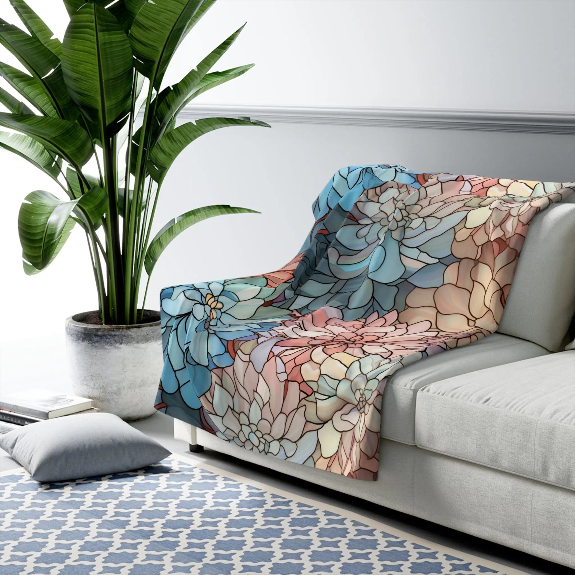 A cozy living room features a white sofa with the Stained Glass Dark Flowers Sherpa Fleece Blanket in blue, pink, and beige. A large green plant sits beside it, a blue and white patterned rug adorns the floor, and nearby is a gray cushion.