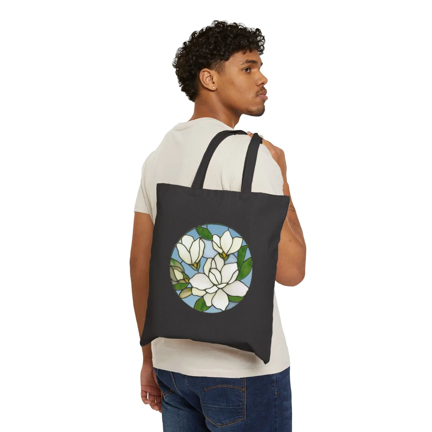 A person in a white t-shirt and jeans holds the black Stained Glass Magnolia Cotton Canvas Tote Bag, featuring white flowers and green leaves on a blue background.