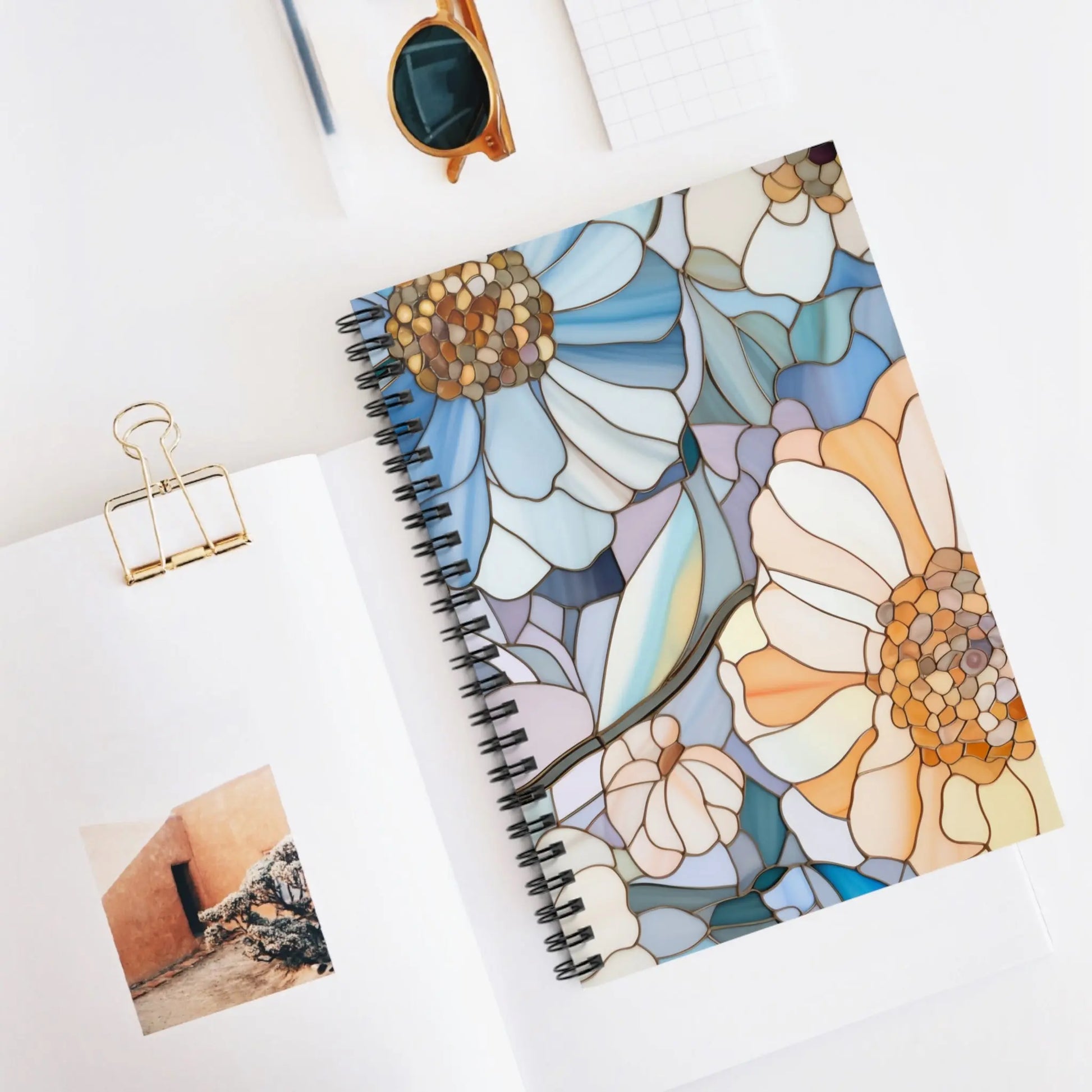The Stained Glass Flowers Spiral Notebook with bespoke printing rests on a white surface. Nearby are an open notebook displaying a small photo, a gold paper clip, square paper, sunglasses, and a pencil.