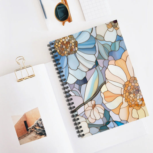 The Stained Glass Flowers Spiral Notebook with bespoke printing rests on a white surface. Nearby are an open notebook displaying a small photo, a gold paper clip, square paper, sunglasses, and a pencil.