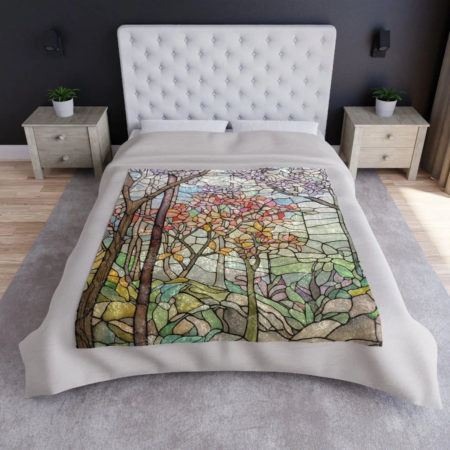 The bed has a white tufted headboard, with two wooden nightstands and potted plants. A custom-made Stained Glass Springtime Forest Crushed Velvet Blanket with a stained glass-style design of fall trees adorns the bed. The gray walls and hardwood floor complete the cozy room ambiance.