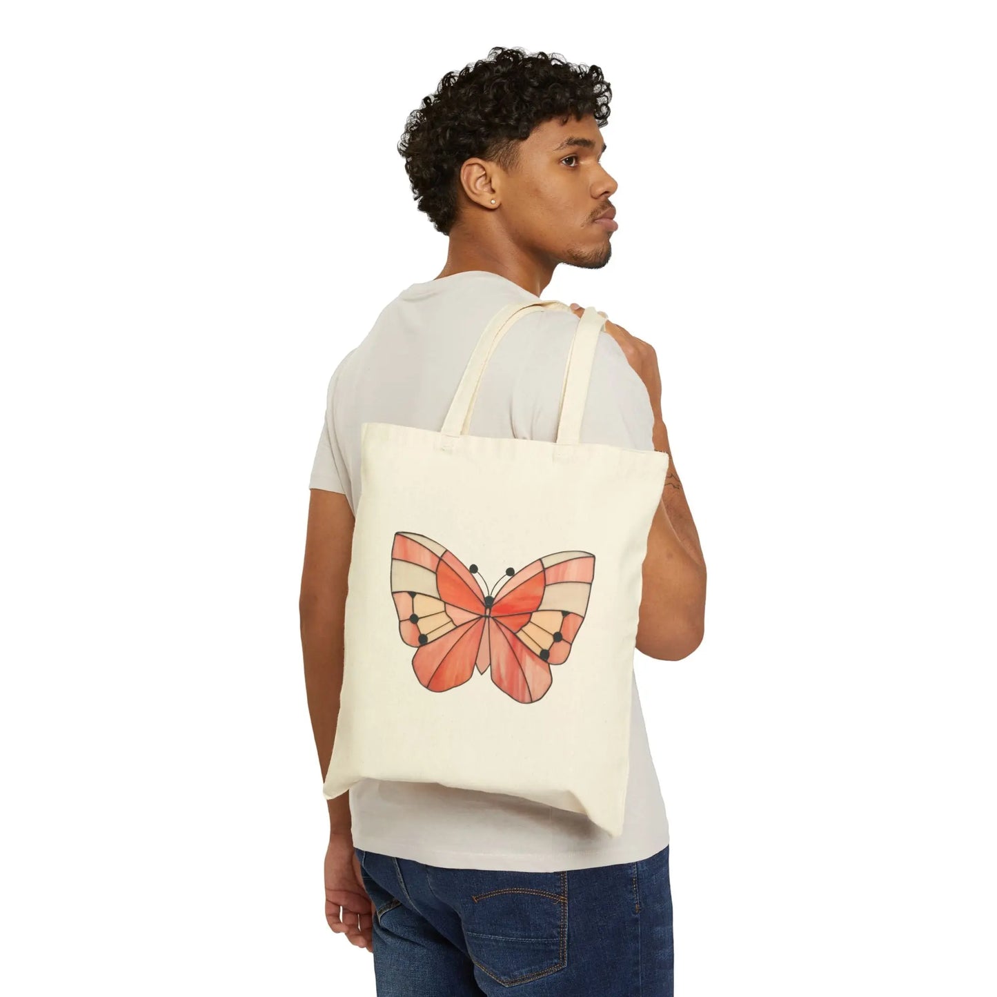 A person with curly hair holds a Stained Glass Spring Butterfly Cotton Canvas Tote Bag over their shoulder, featuring vibrant butterfly art. Theyre in a light t-shirt and blue jeans, set against a white backdrop.