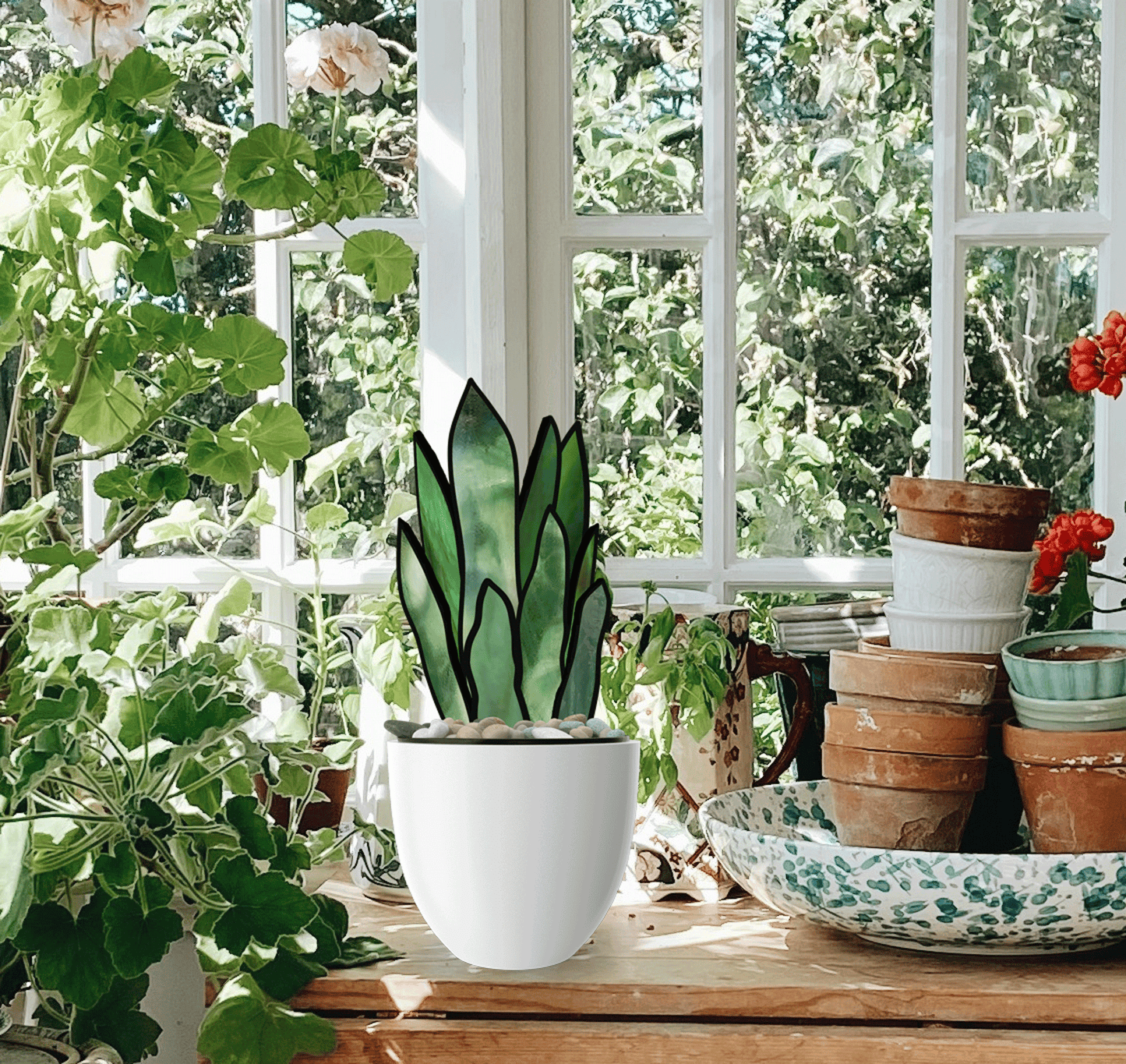 A white pot with a plant design is on a wooden table, surrounded by clay pots and a decorative plate. In the sunlit room, greenery and flowers cast vibrant shadows that resemble the 3D Snake Plant Leaves Stained Glass Pattern through the window.