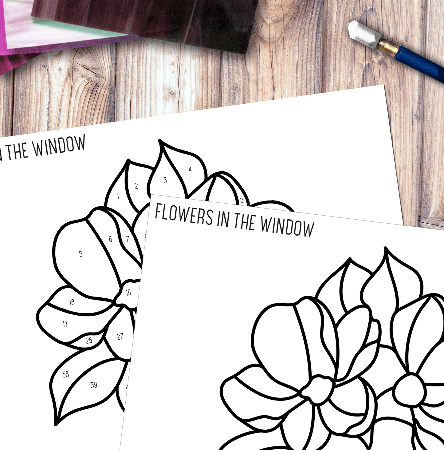 Two sheets titled Bundle of Flowers Stained Glass Pattern rest on a wooden table with a pencil nearby, featuring large outlined flowers like stained glass, each section numbered for vibrant coloring as if creating a suncatcher.