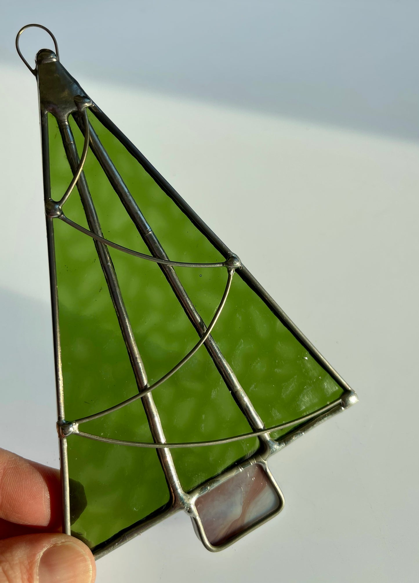 A hand holds a Handmade Christmas Tree Suncatcher by Noelle Barnes, crafted in the Tiffany-method. The ornament features triangular green glass pieces outlined in metal on a light rectangular base, softly illuminated from the right.