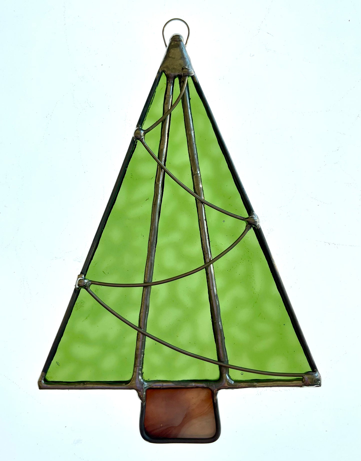 The Handmade Christmas Tree Suncatcher by Noelle Barnes features vibrant green glass panels and a brown trunk outlined in metal. This charming ornament, crafted with the Tiffany-method stained glass technique, includes a metal wire loop for easy hanging.