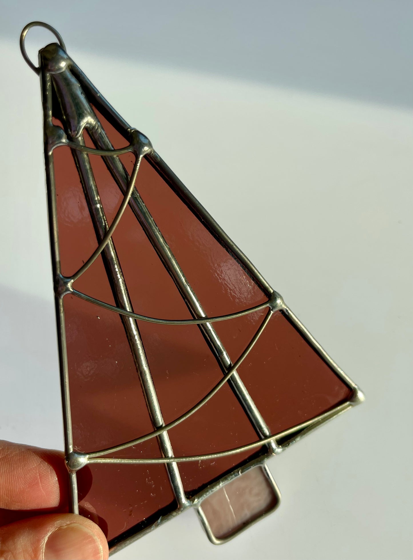 A hand holds a Handmade Christmas Tree Suncatcher by Noelle Barnes, featuring Tiffany-method stained glass in a rich brown hue. Silver soldering forms swirling patterns on this slightly translucent ornament, which beautifully captures light.