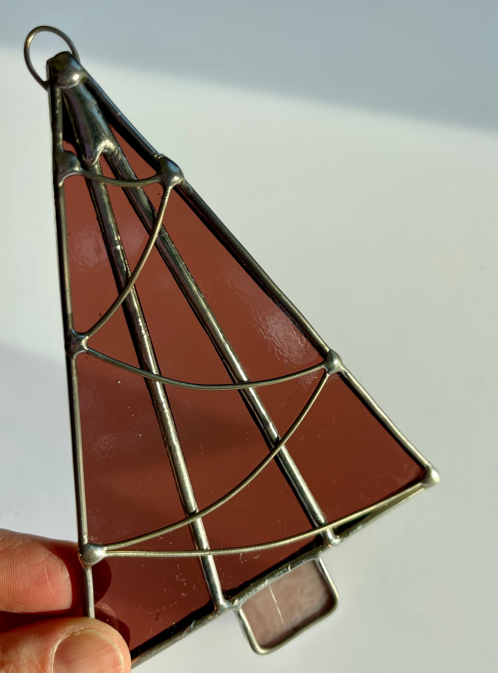 A hand holds a Handmade Christmas Tree Suncatcher by Noelle Barnes, featuring Tiffany-method stained glass in a rich brown hue. Silver soldering forms swirling patterns on this slightly translucent ornament, which beautifully captures light.