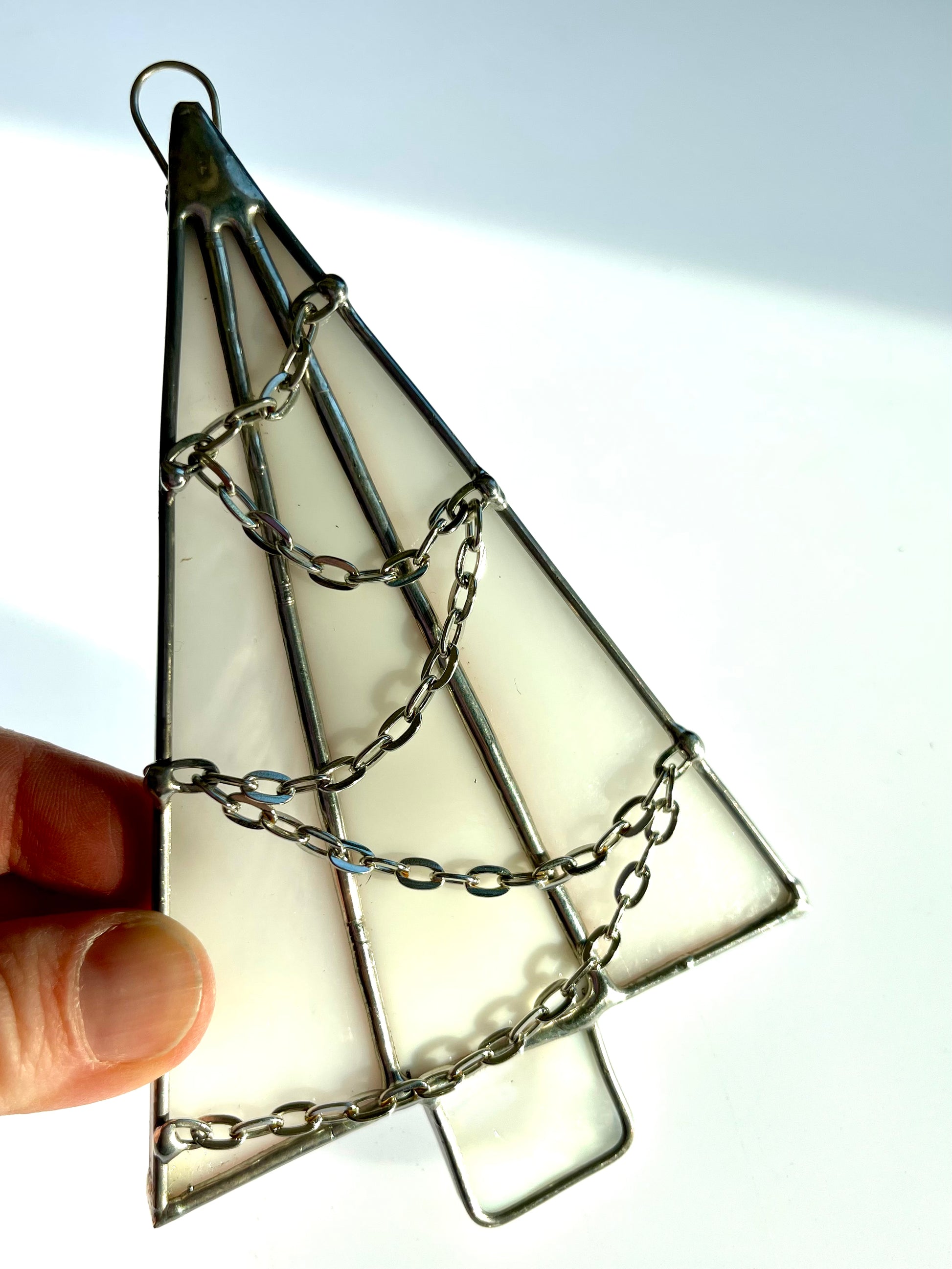 A hand holds a Handmade Christmas Tree Suncatcher by Noelle Barnes, featuring three triangular white glass panels with silver edges and a draped chain. The softly lit background enhances its festive charm.