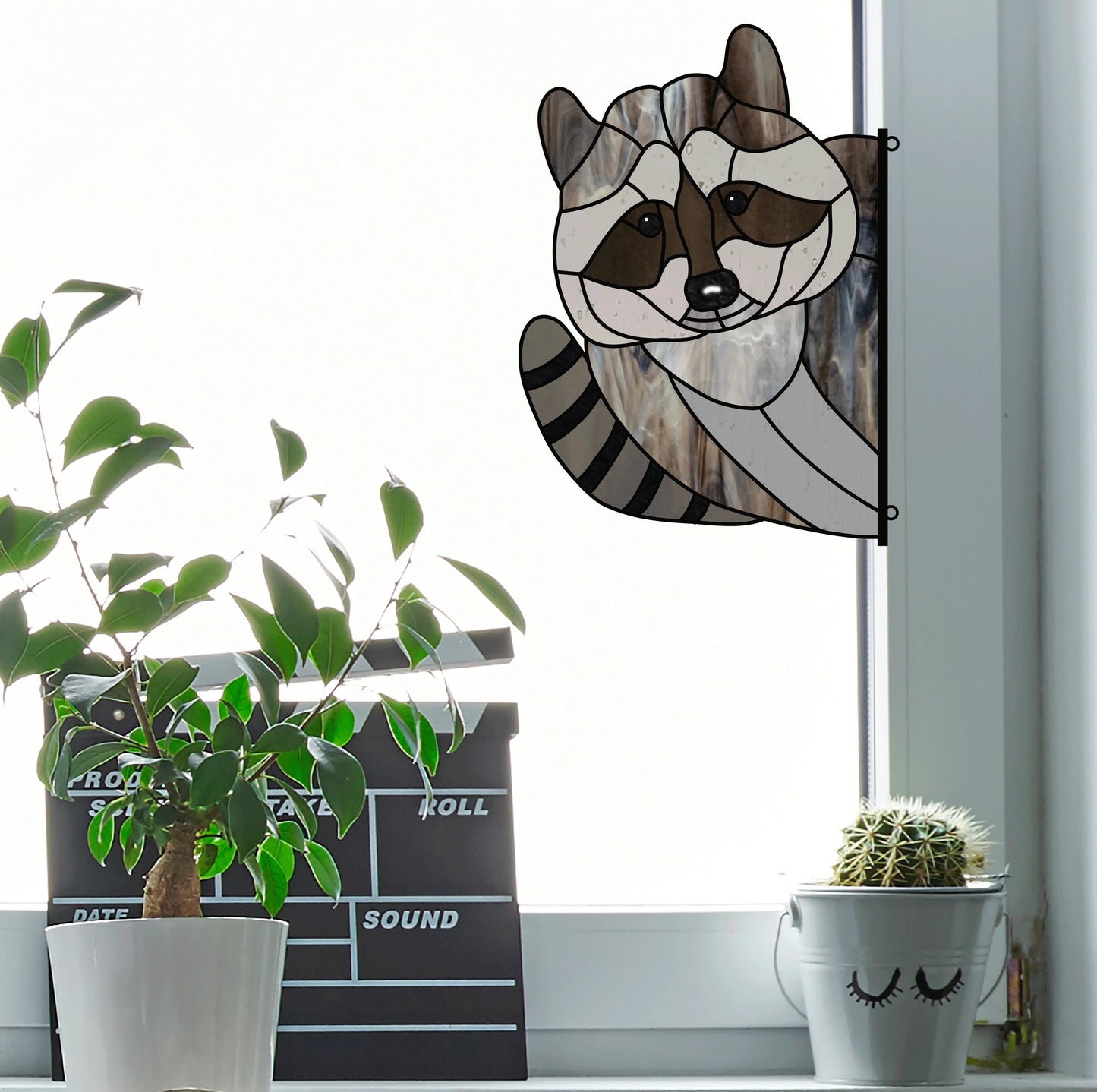 An Animal Stained Glass Pattern from the 9-Pack hangs on a window, casting digital-like patterns. Below, a potted plant and cactus with a face design sit on the sill. A clapperboard rests to their left, while bright, soft light enhances this intermediate skill level scene.