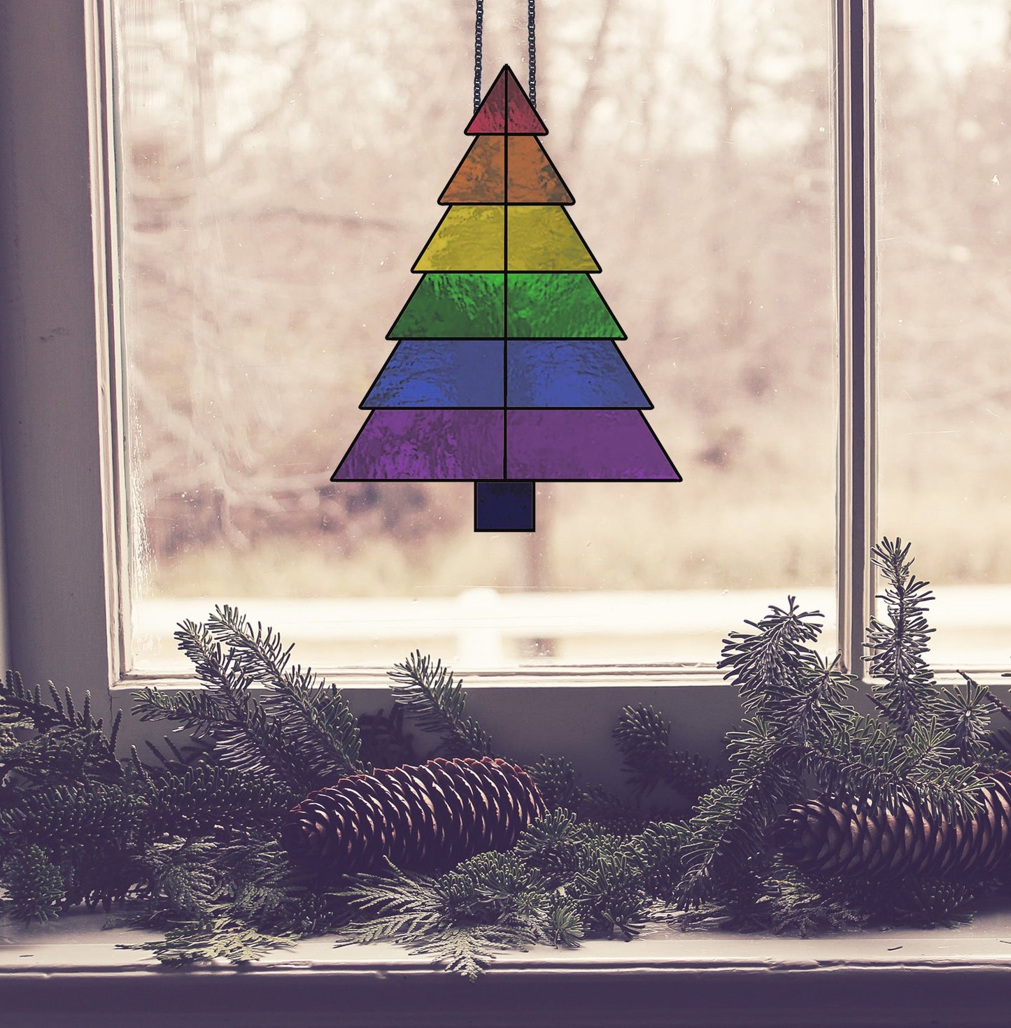 A Beginner Christmas Tree Rainbow Stained Glass Pattern suncatcher hangs in a window, framed by evergreen branches and pinecones on the sill, with a blurred outdoor scene beyond.