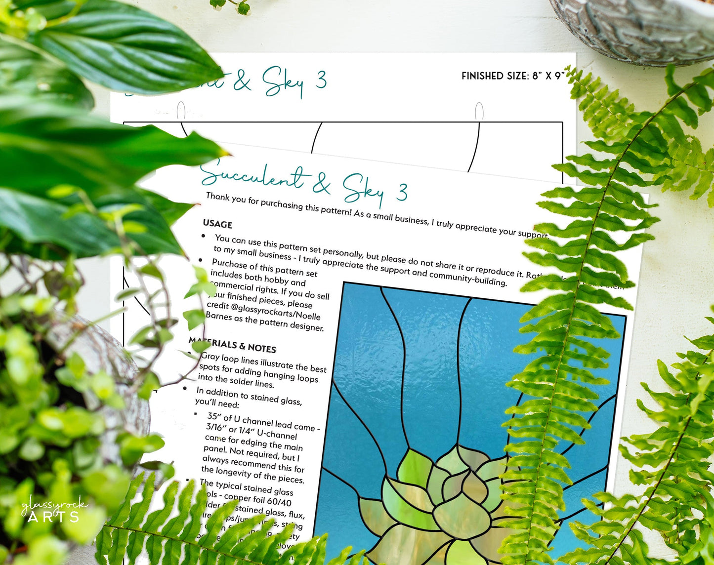 The Stained Glass Succulent Panel Pattern lies on a surface surrounded by green ferns. This digital design comes with usage instructions, general notes, and an option for a commercial license for creative projects.
