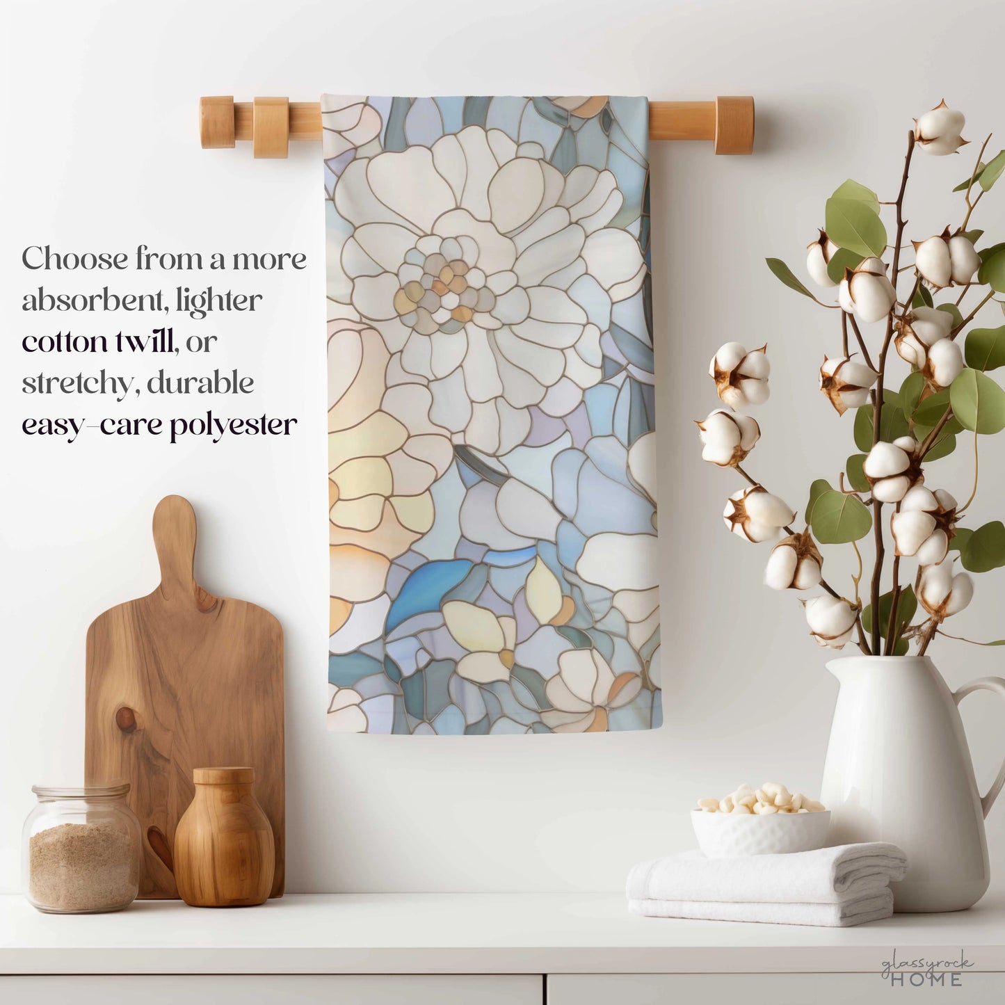 The Stained Glass Flowers Kitchen Towel hangs on a wooden holder, enhancing the kitchen decor. Nearby, a cotton stem vase, cutting board, grain jars, rolled towel, and potted plant complete the scene. Text promotes choice of absorbent cotton twill or stretchy polyester material.