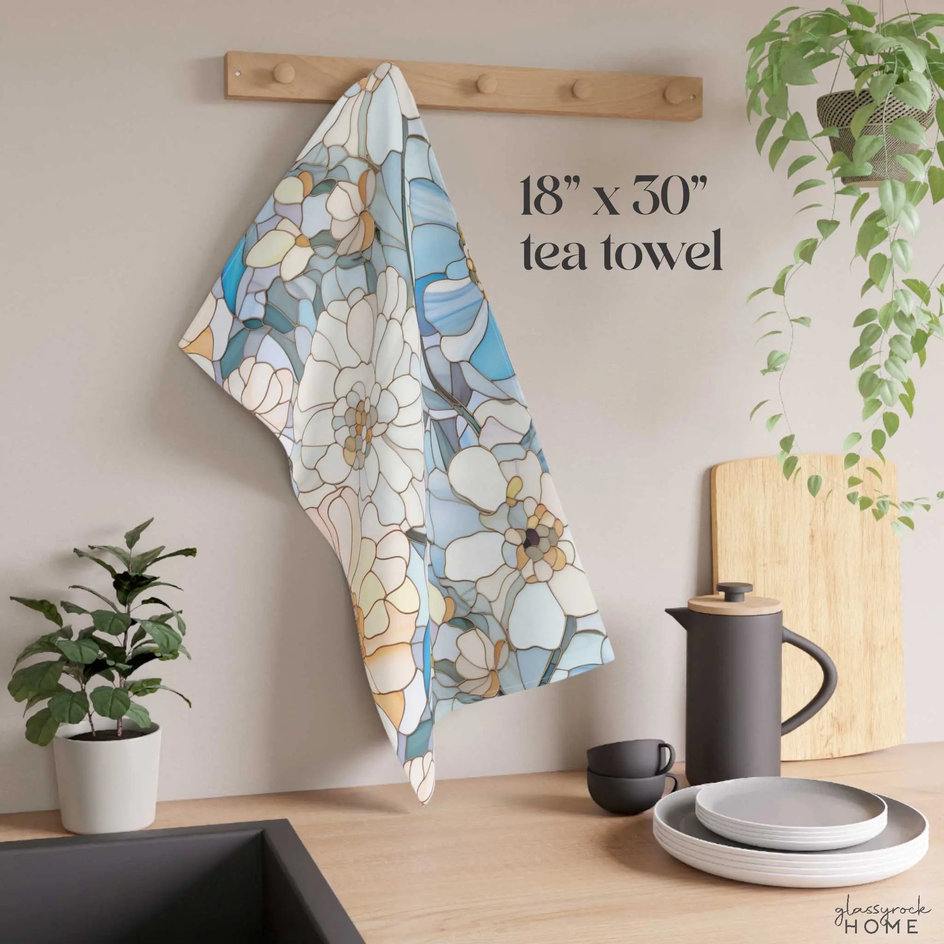 The Stained Glass Flowers Kitchen Towel, with a floral design in blue, white, and beige, hangs on a wooden hook, enhancing the kitchens look. Below sits a counter with stacked plates, a black teapot and cup, cutting board, and potted plant. Text reads 18 x 30 tea towel.