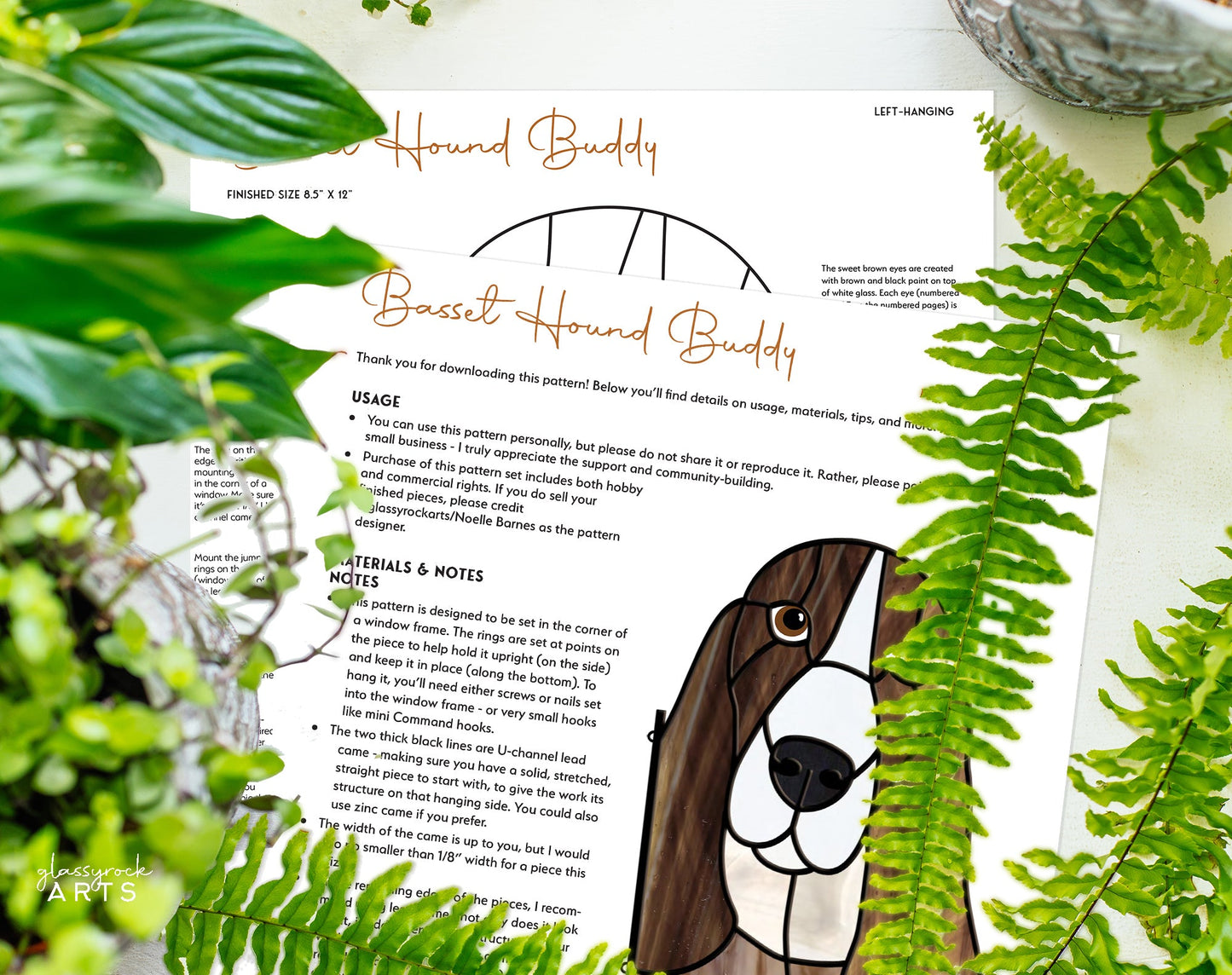 A pattern titled Basset Hound Dog Stained Glass Pattern rests on a surface amid lush greenery, featuring instructions and a stylized image of a Basset Hounds head, reminiscent of charming stained glass art.