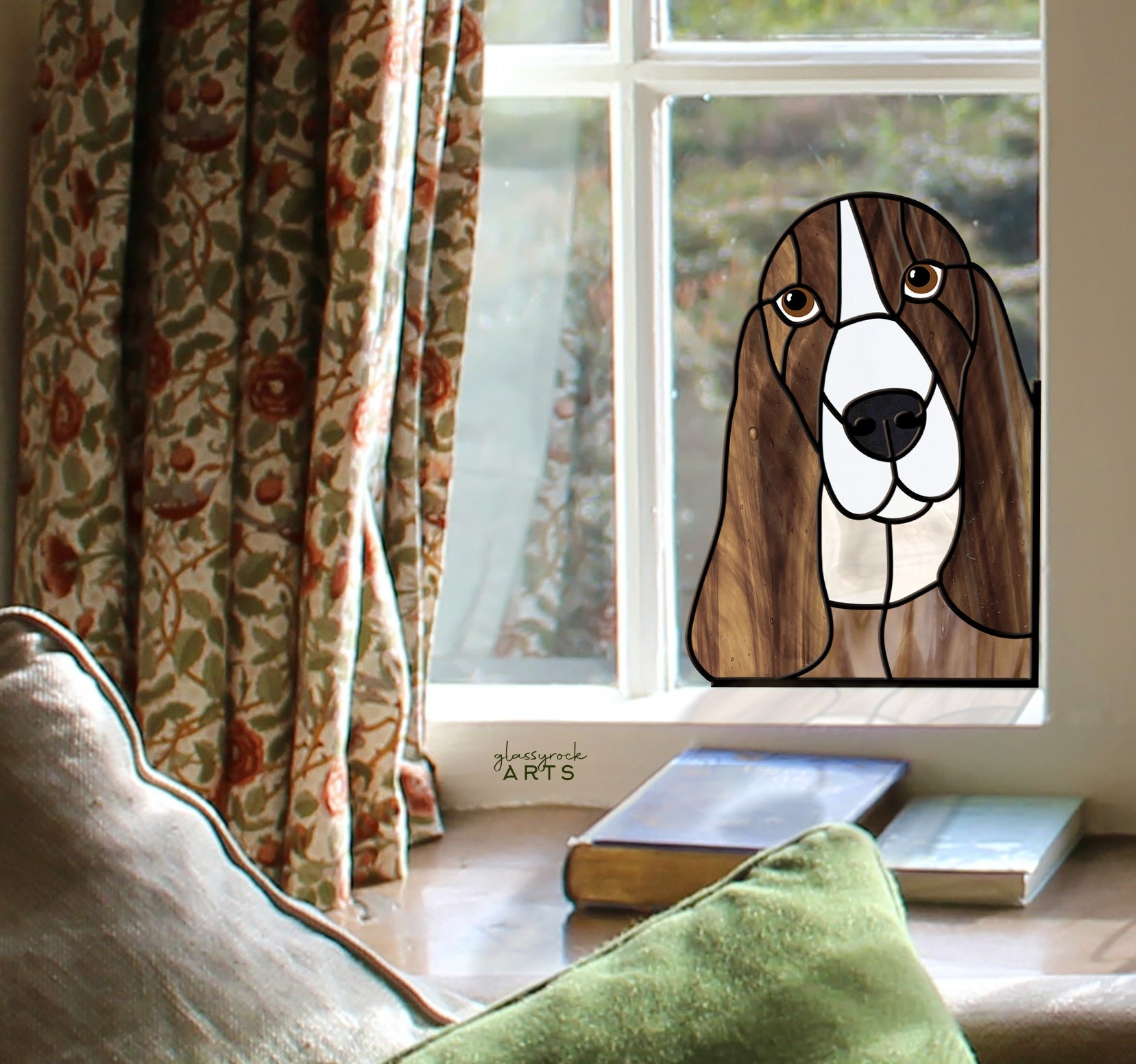 A Basset Hound Dog Stained Glass Pattern serves as an elegant suncatcher in the window. Framed by floral curtains, sunlight floods the room while stacked books and a patterned cushion beckon you to unwind on the cozy chair.