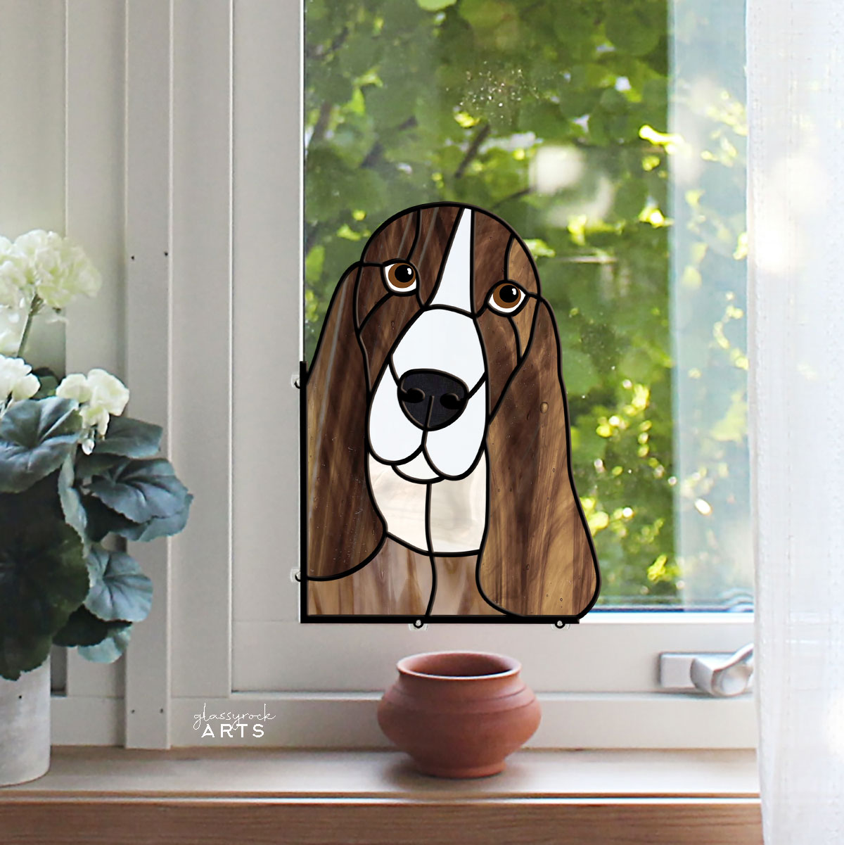 The Basset Hound Dog Stained Glass Pattern features a brown and white Basset Hound with long ears in a sunlit window, complemented by white flowers and a small brown pot on the sill. Green foliage outside adds to the shimmer of the intricate design.