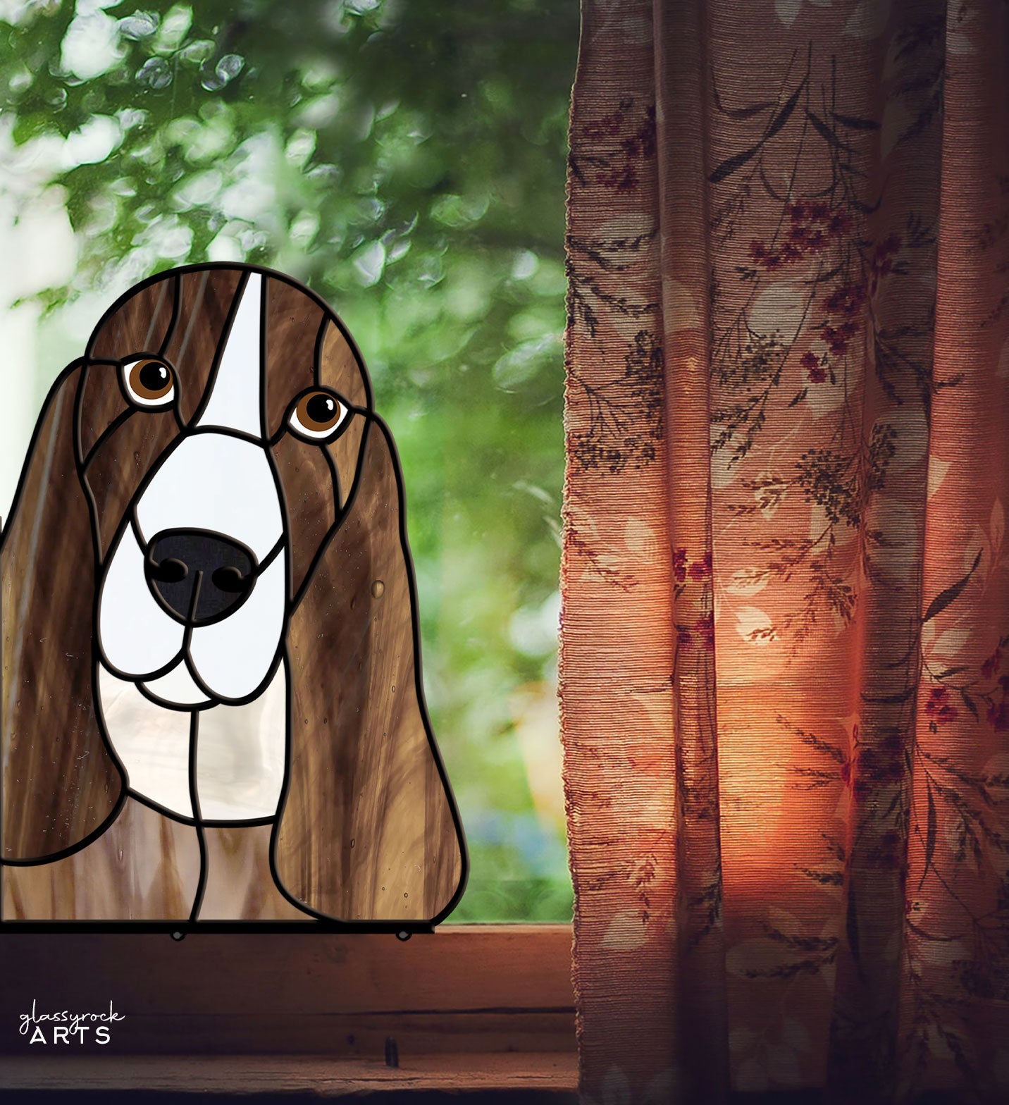 A Basset Hound Dog stained glass artwork rests on a windowsill. A floral curtain is partly drawn, letting soft light shine through, illuminating the Basset Hound art. Lush greenery can be seen outside the window.