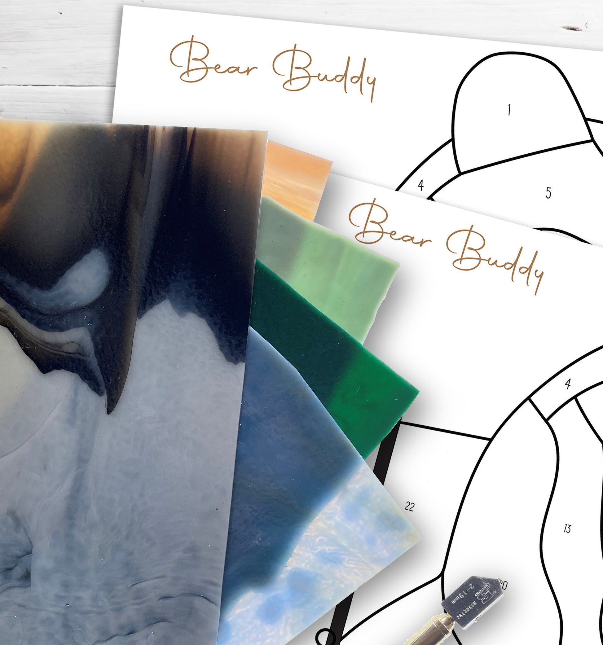 Four sheets of stained glass in various colors overlap two Big Bear Buddy Stained Glass Pattern paper patterns. A glass cutter rests nearby on the white surface, with an option for a digital pattern download available.