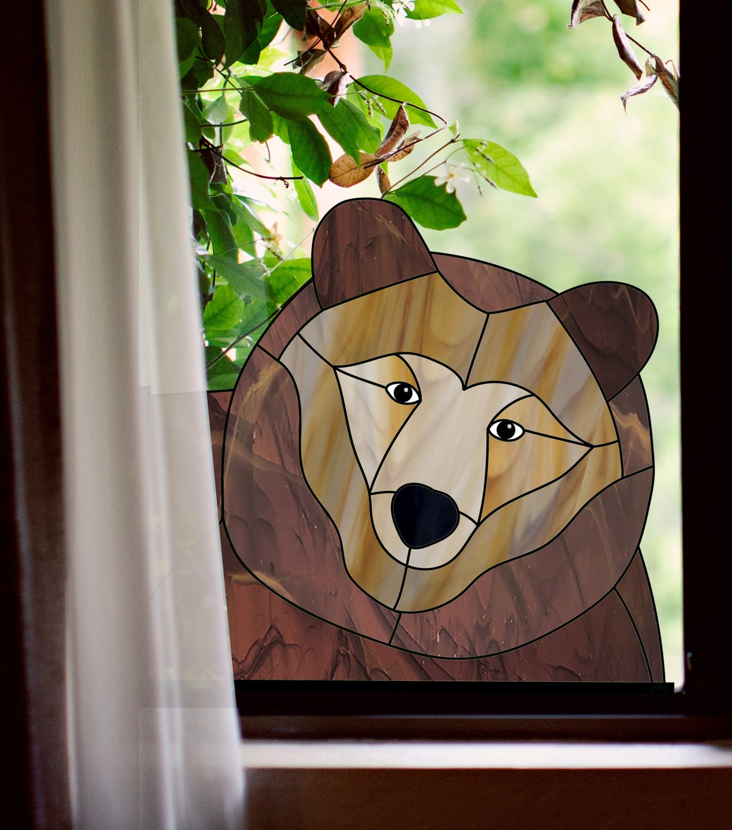 The Big Bear Buddy Stained Glass Pattern, with brown and beige tones, is displayed near a window. Sunlight filtering through the trees provides a natural backdrop for this exquisite glass art piece resembling a bears face.