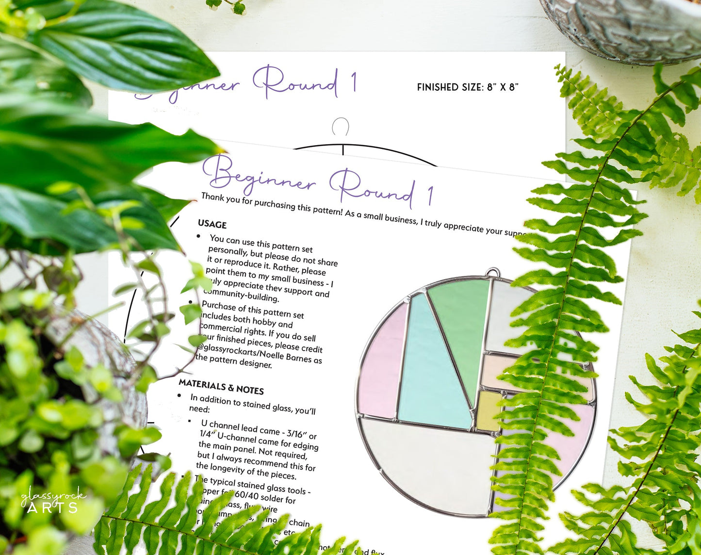 A Round Beginner Stained Glass Pattern instruction sheet is surrounded by green ferns with a partially assembled circular piece featuring pastel sections and geometric patterns on top.