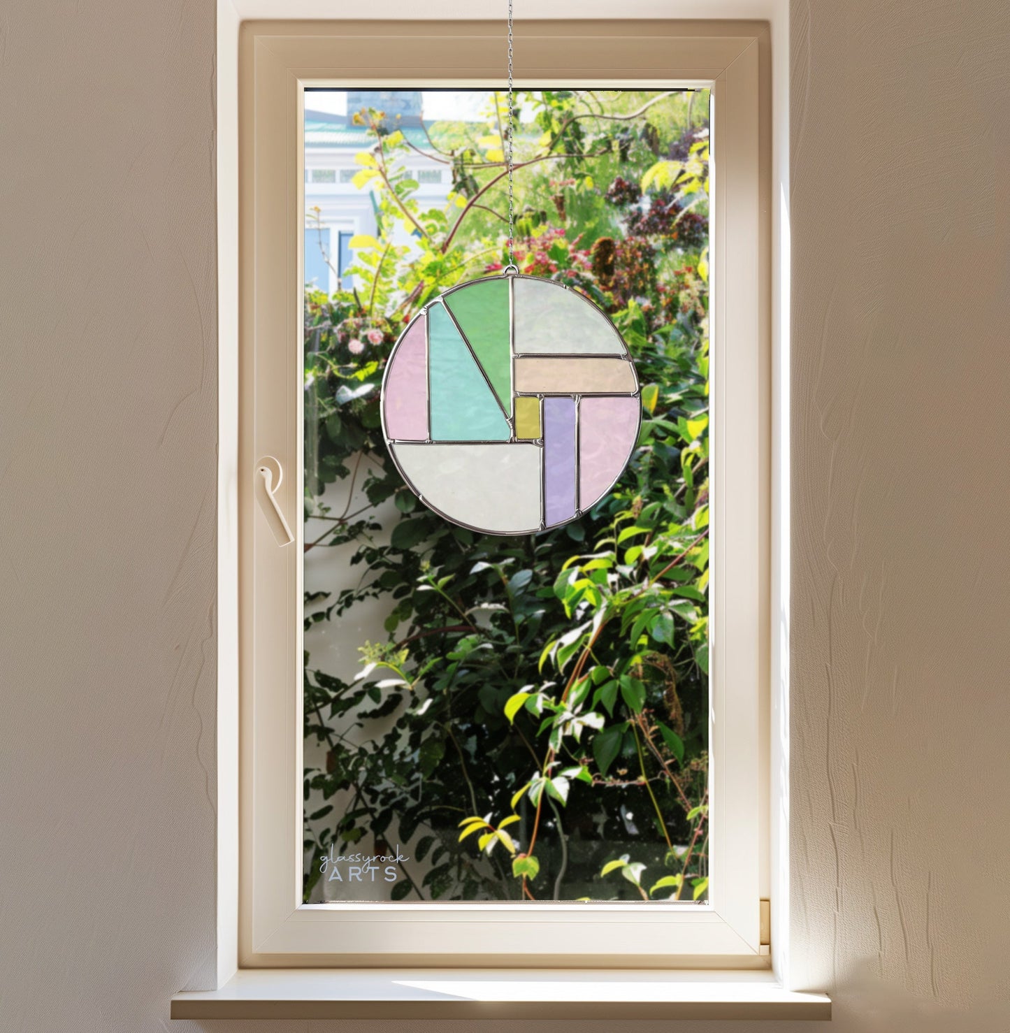 The Round Beginner Stained Glass Pattern features a captivating geometric design in pastel hues, hanging in a window. Sunlight cascades through, illuminating the lush green garden outside. The window is nestled within a beige wall framed by simple white trim.