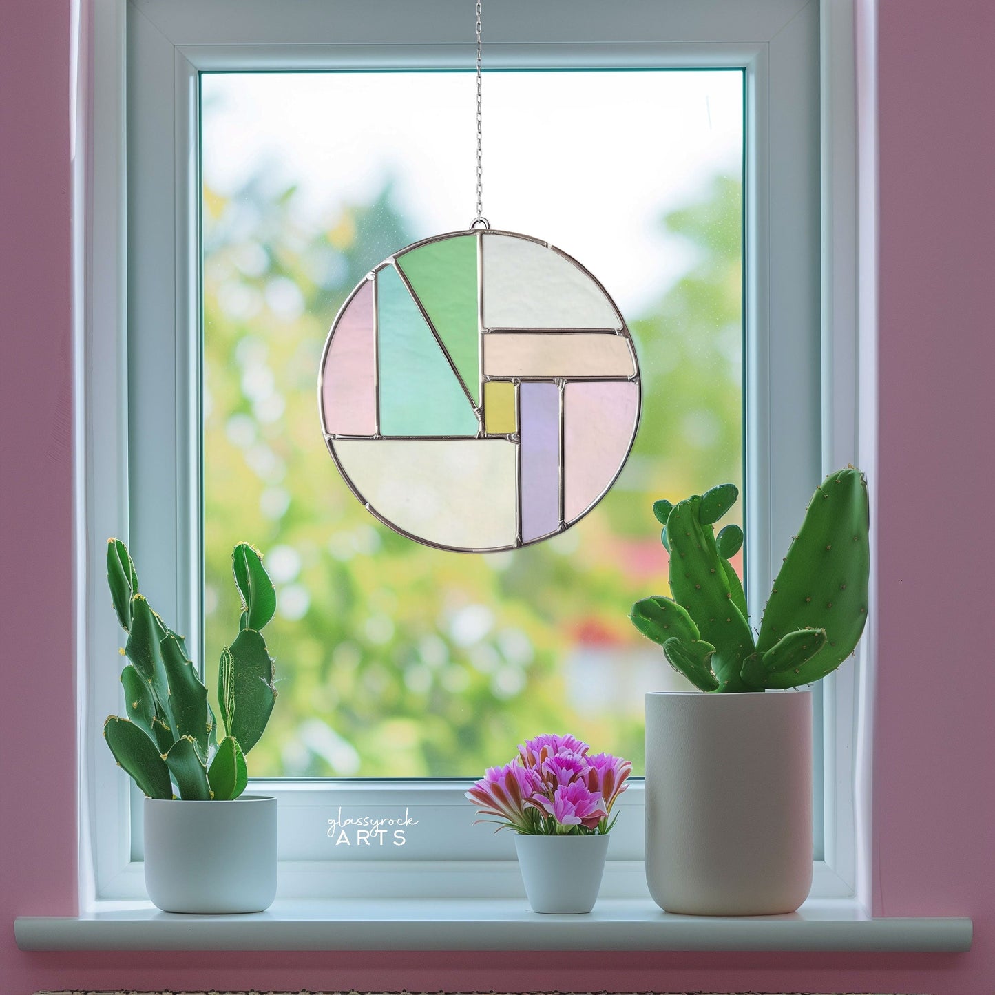 A Round Beginner Stained Glass Pattern hangs in the window, bordered by two cacti and a small pot of purple flowers on the sill, with a blurred view of greenery outside.