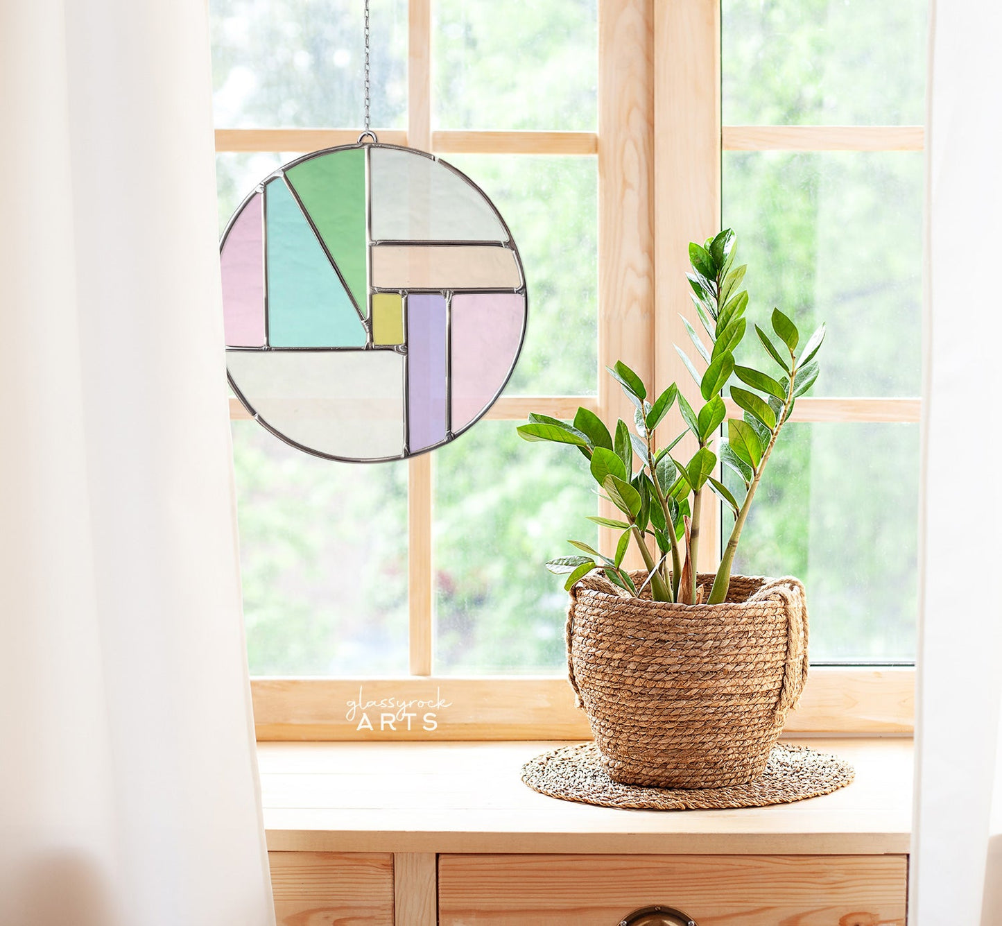 A Round Beginner Stained Glass Pattern with a vibrant geometric design hangs in a bright window. Below, a green leafy plant sits in a woven basket on a wooden table. White curtains frame the scene elegantly, revealing lush greenery outside.