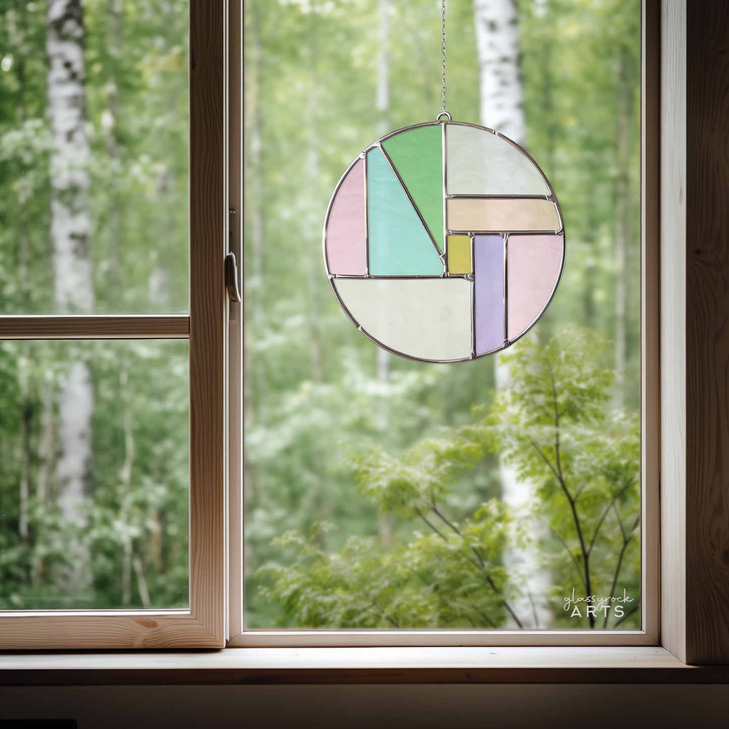 The Round Beginner Stained Glass Pattern, featuring geometric pastel shapes and a Fibonacci spiral, hangs in a wooden frame, beautifully showcasing a lush birch-dotted forest. Available as a digital pattern download.