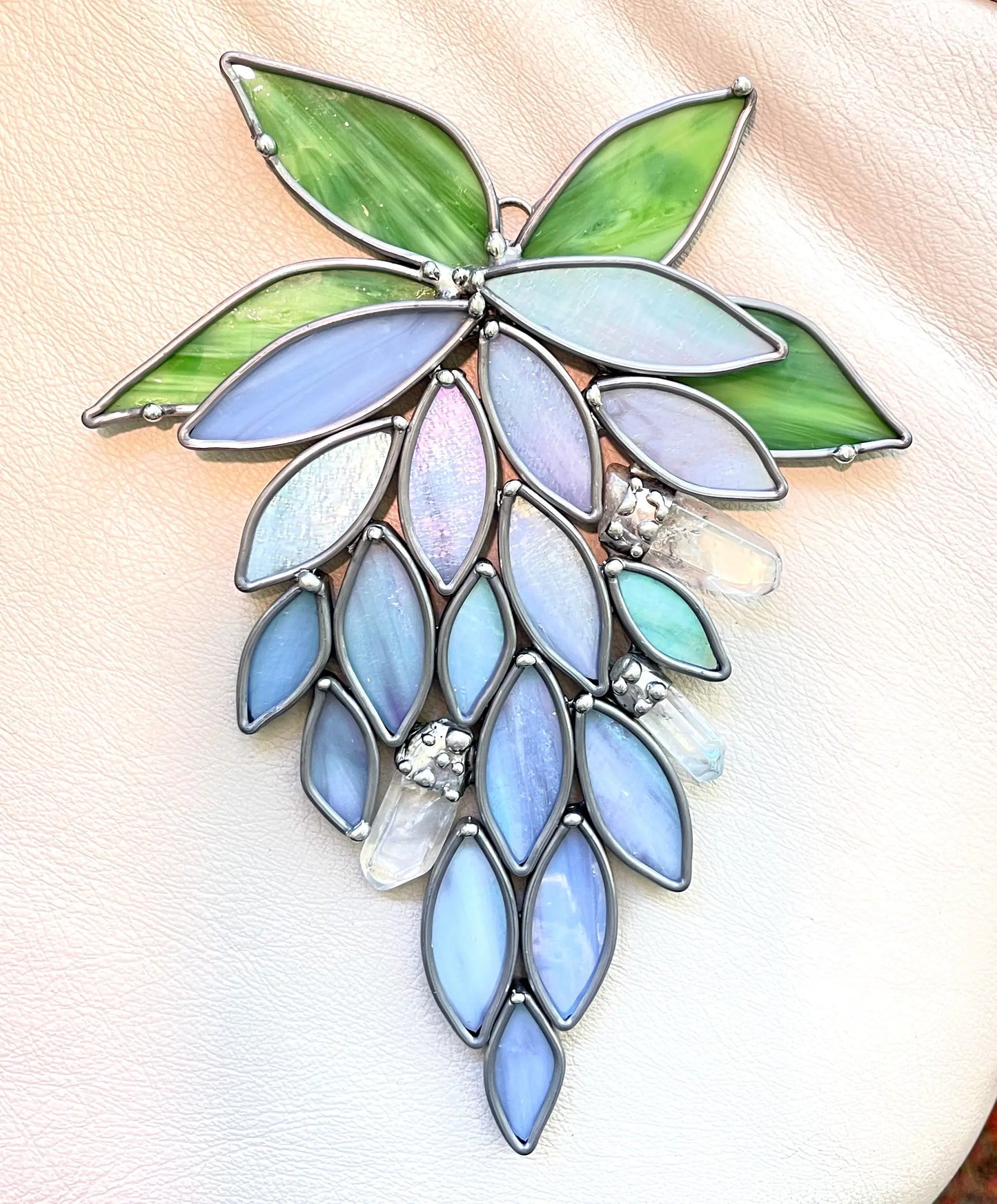 The Handmade Stained Glass Crystal Wisteria Suncatcher showcases blue and purple grape clusters with vibrant green leaves on a light beige fabric, sparkling like crystals in sunlight.