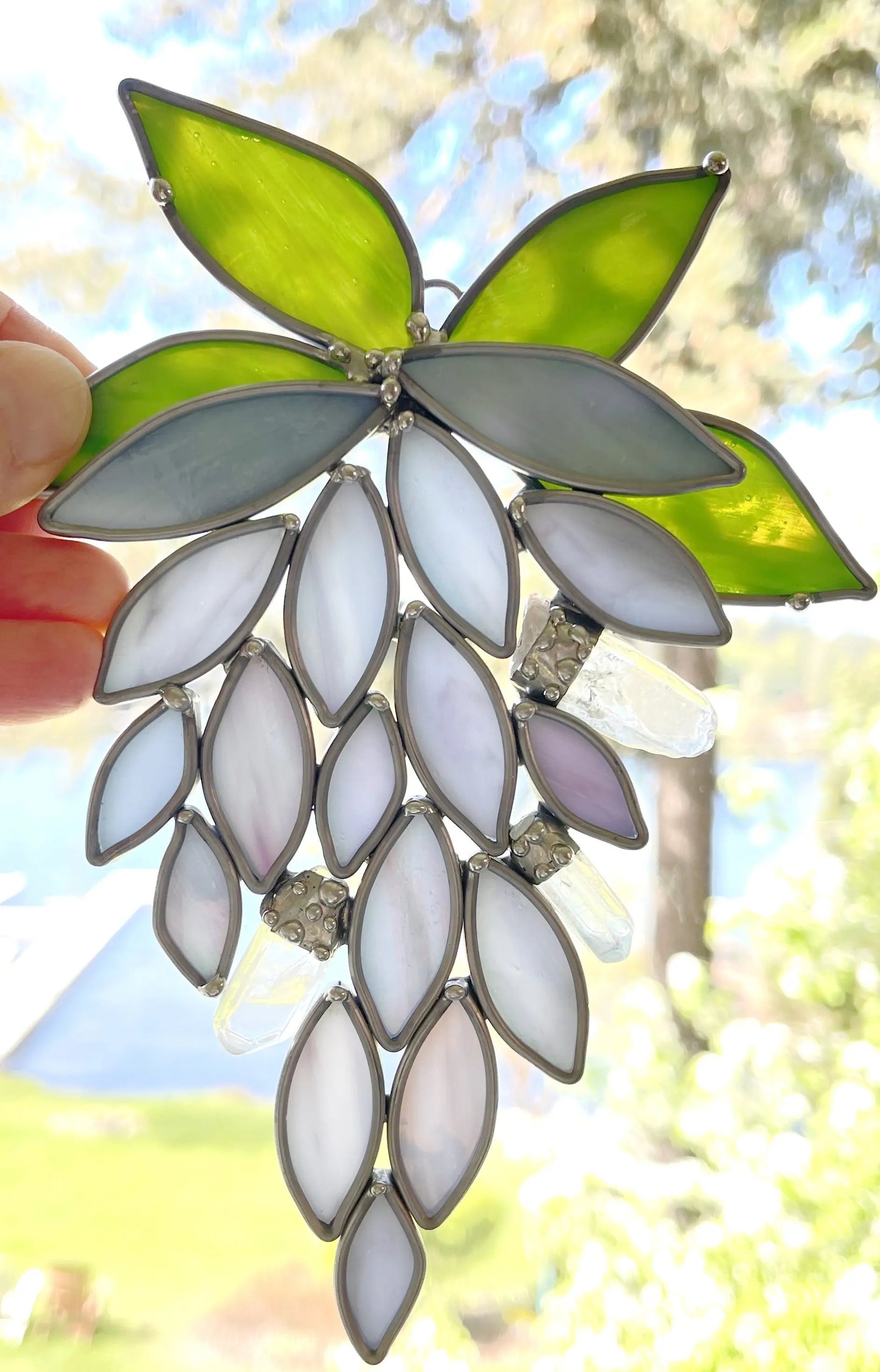 A hand holds a Handmade Stained Glass Crystal Wisteria Suncatcher with purple and white grapes and green leaves against a blurred tree and water background, as sunlight streams through, casting vibrant colors like precious crystals.