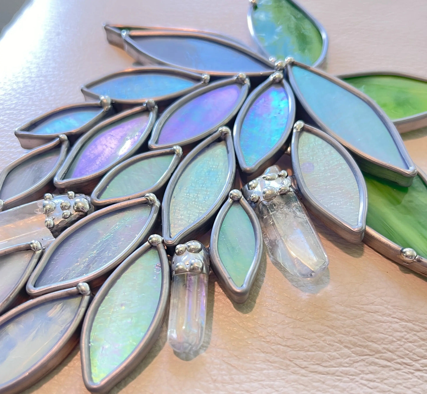 The Handmade Stained Glass Crystal Wisteria Suncatcher features a leaf-like design with blue and green iridescent pieces, accented by two clear crystals and elegantly framed in metal, creating a dazzling effect against light.