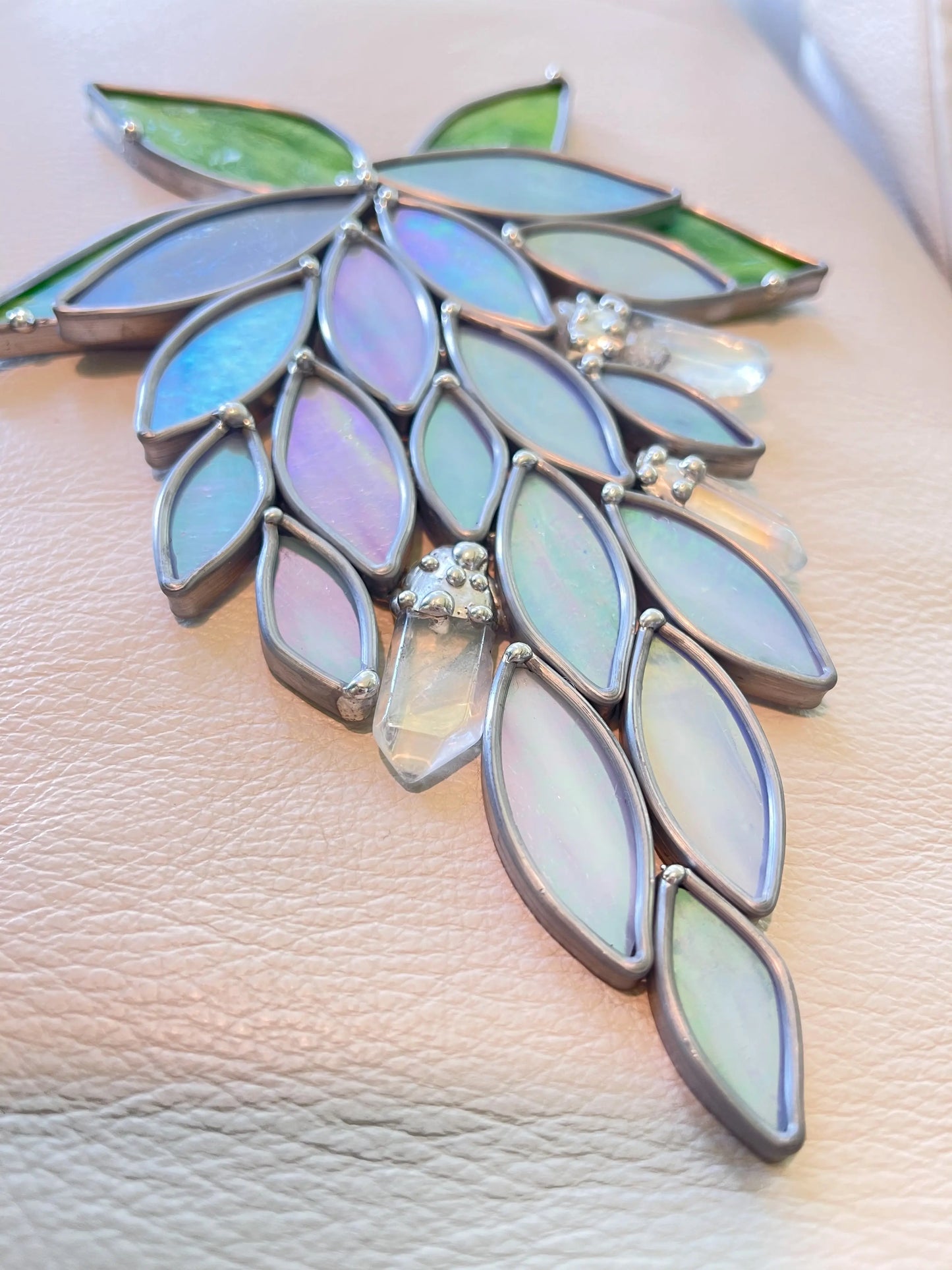 The Handmade Stained Glass Crystal Wisteria Suncatcher showcases leaf-shaped glass in blue, purple, and green with metal settings. It evokes a cascading floral design elegantly placed on soft beige leather.